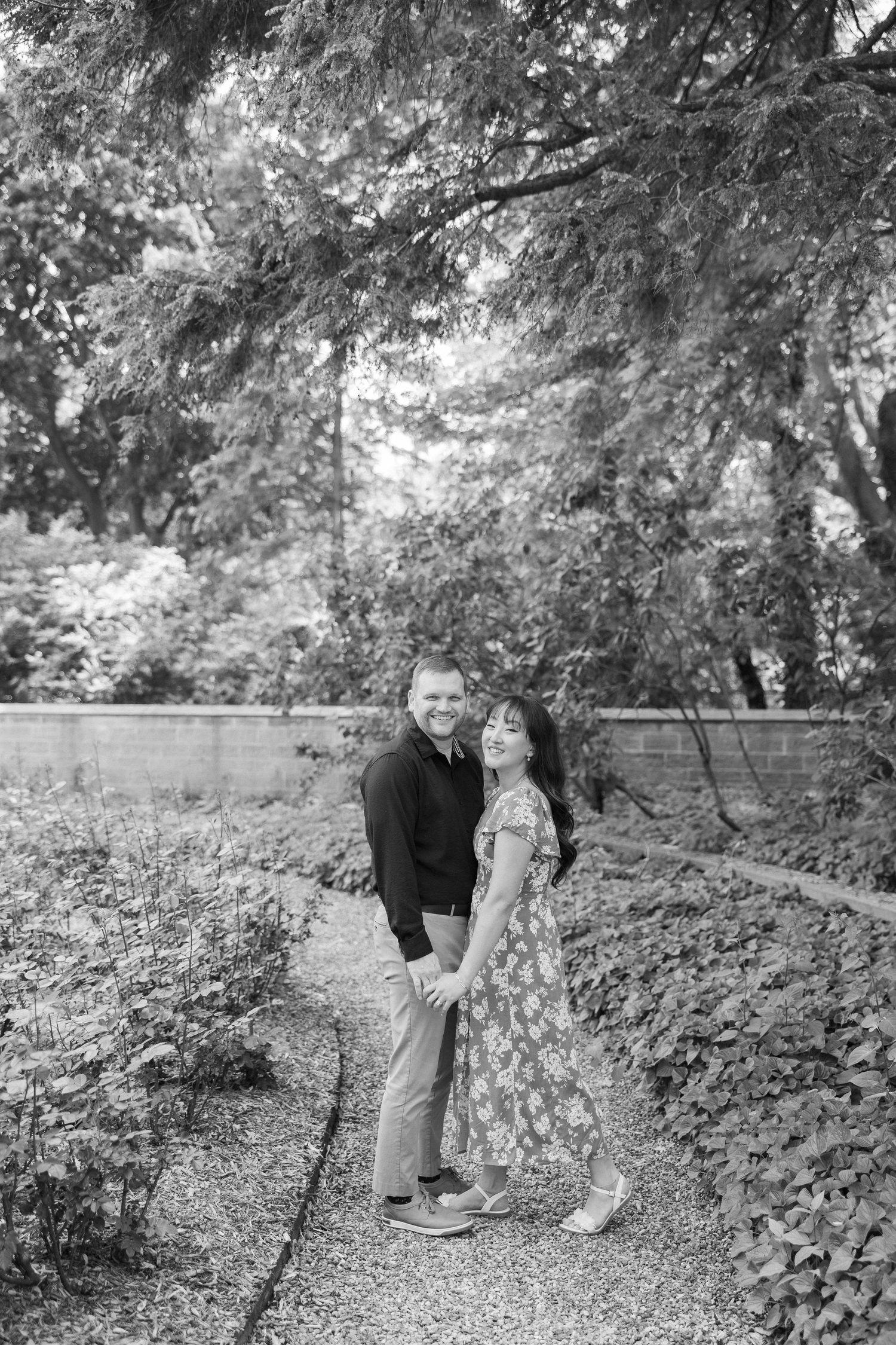 Black and white portrait of couple standing together on garden path under trees.