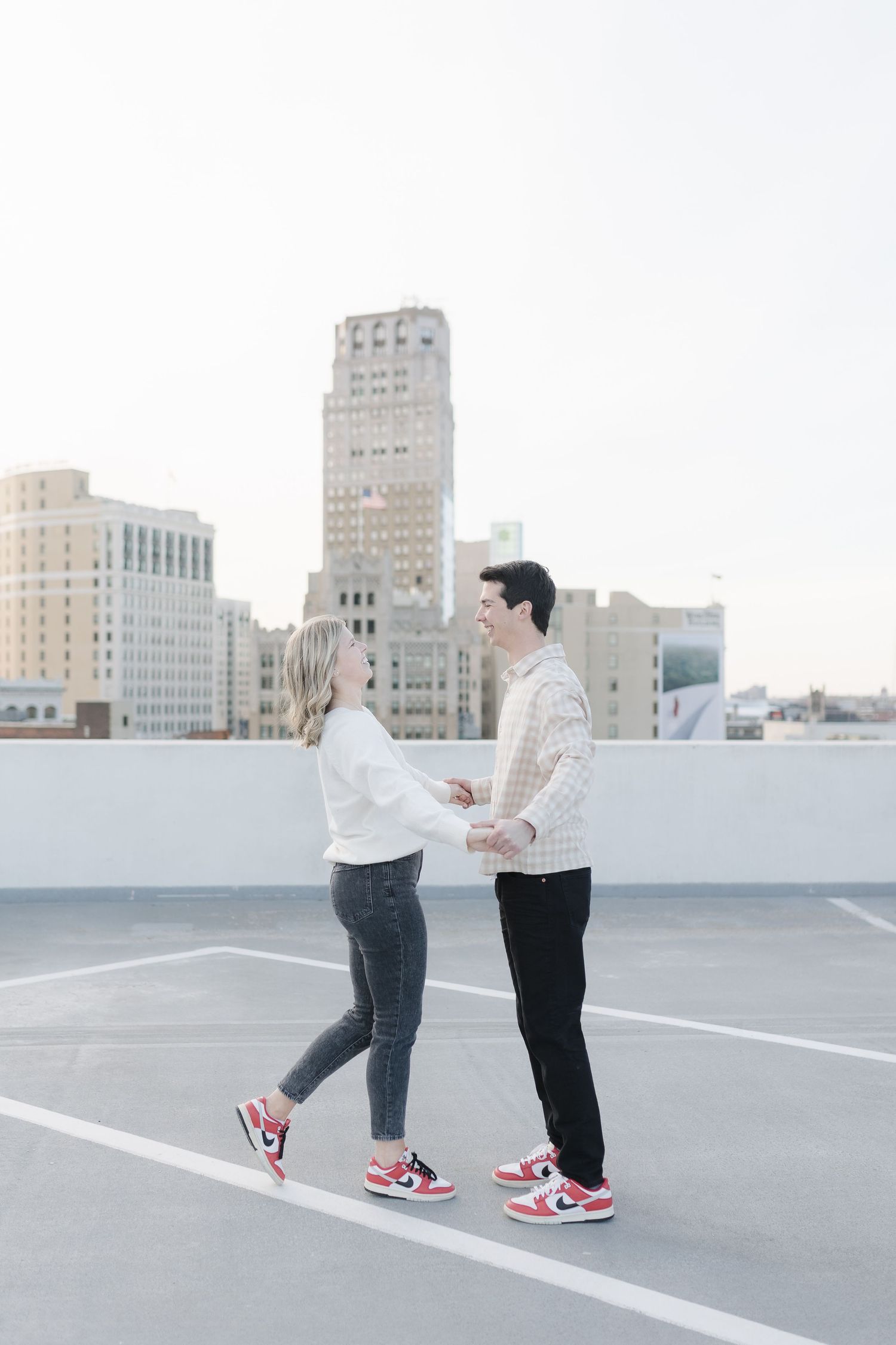 A couple wearing matching white sweaters share a romantic moment on a downtown rooftop parking deck.