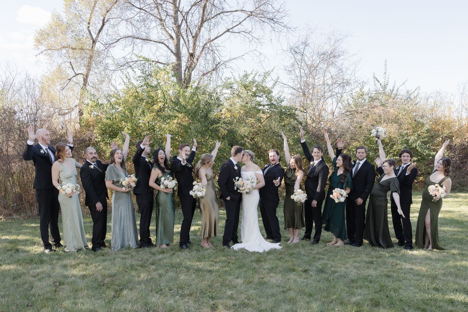 A wedding party in formal attire stands outdoors on grass with bare trees and greenery in the background.