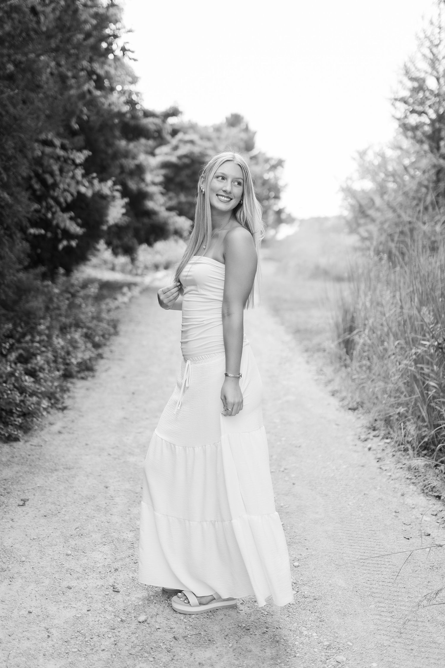 A woman in a flowing white strapless dress poses on a rural dirt path surrounded by greenery in black and white.