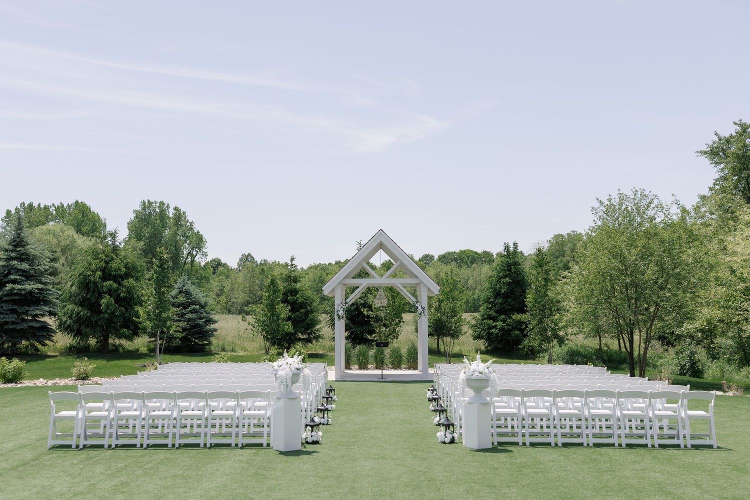 Elegant outdoor wedding ceremony setup with white chairs, floral arrangements, and a geometric arch on a lush green lawn.