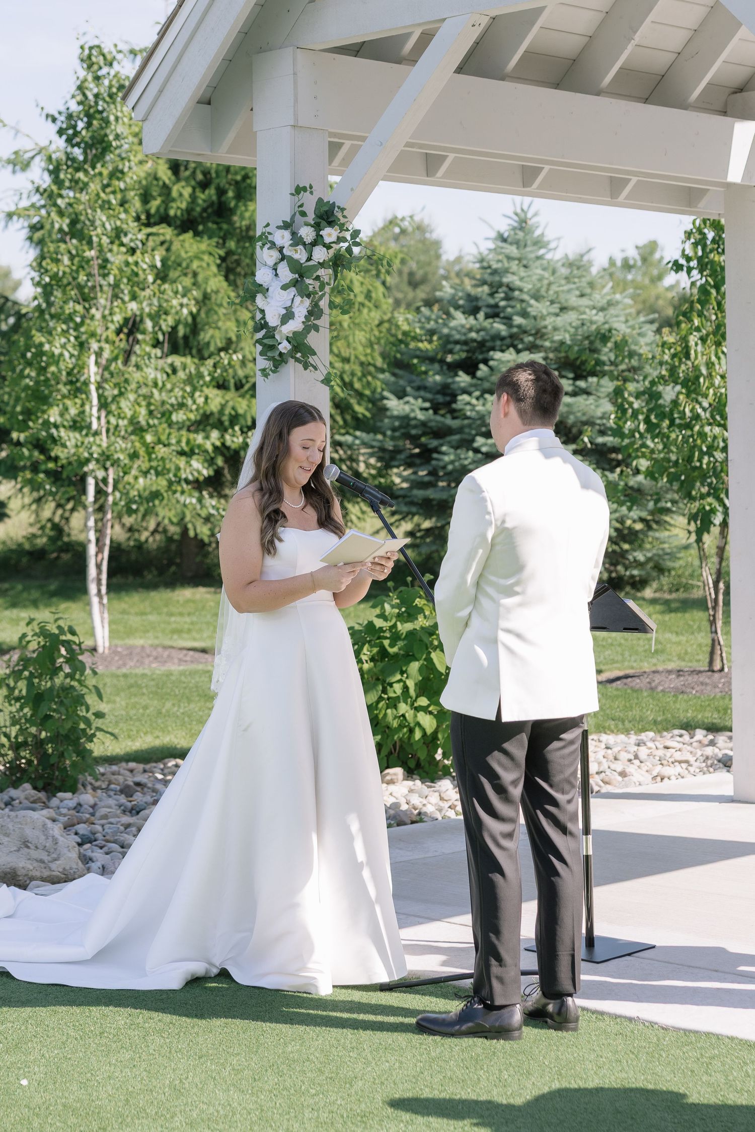 Bride reads wedding vows to groom under white outdoor pavilion surrounded by lush greenery on sunny wedding day.