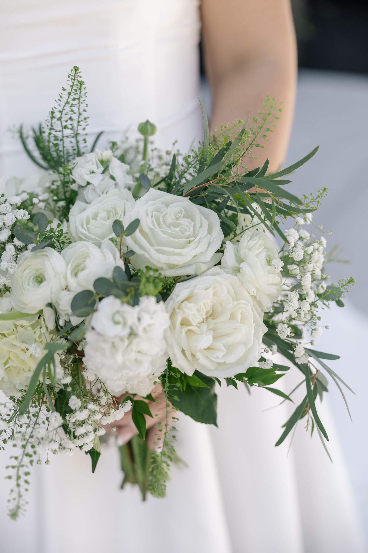 Bride holding a lush white wedding bouquet with roses, ranunculus, baby's breath, and greenery in an elegant arrangement.