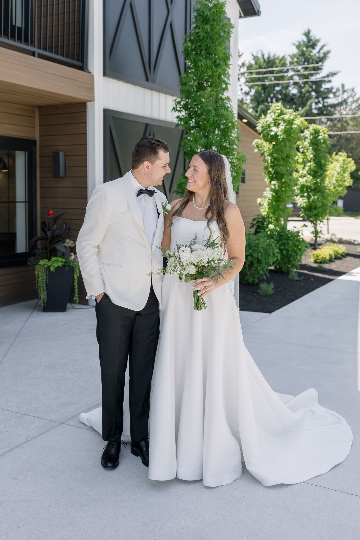 A bride and groom pose together outdoors, she in a white gown with bouquet, he in a white tuxedo jacket with bow tie.