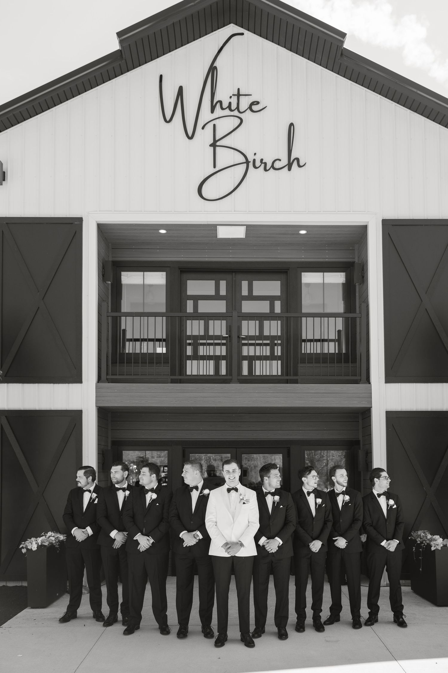 Groom and groomsmen in black tuxedos pose outside White Birch wedding venue in elegant black and white photo.