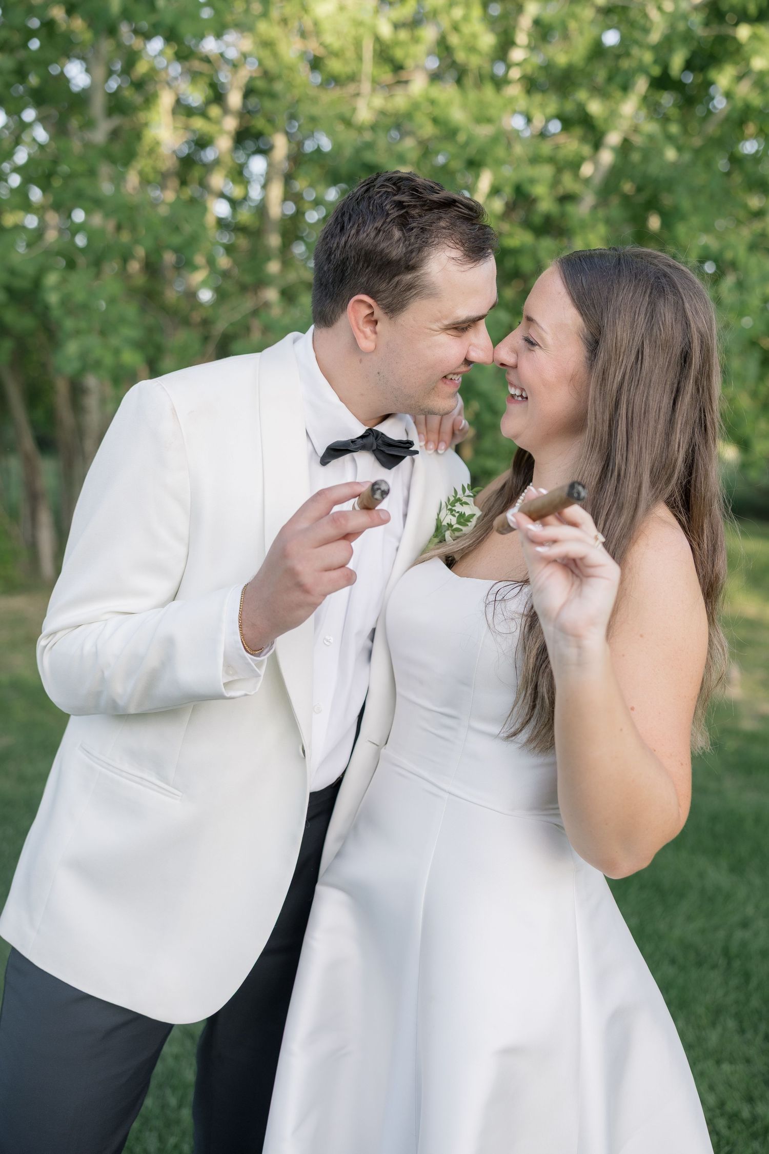 A smiling bride and groom in white wedding attire share a romantic moment while holding cigars outdoors.