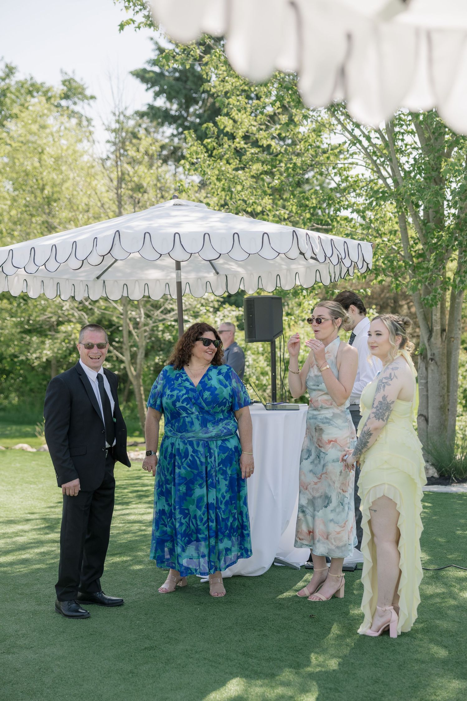 Outdoor wedding cocktail hour guests mingling on green lawn under white scalloped umbrella on sunny day.