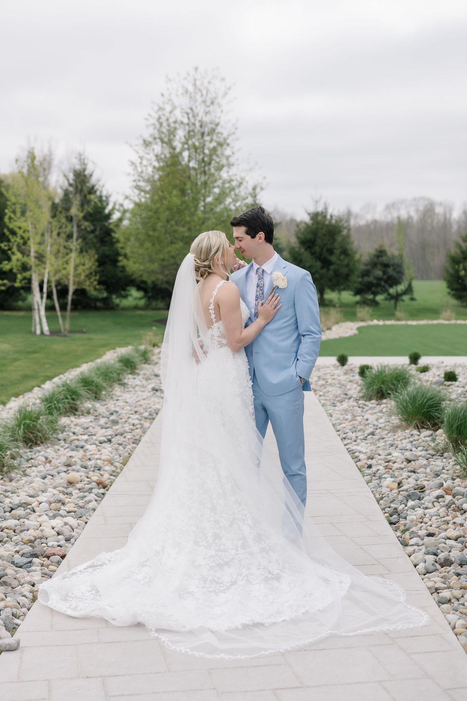 A bride and groom share a romantic moment on a stone path, her lace gown and veil trailing beautifully behind them.