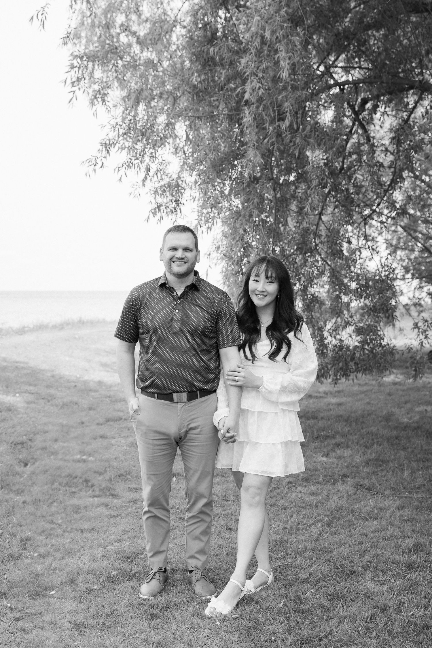 A couple shares a joyful moment together on a grassy hill under willow trees in a romantic outdoor photoshoot.