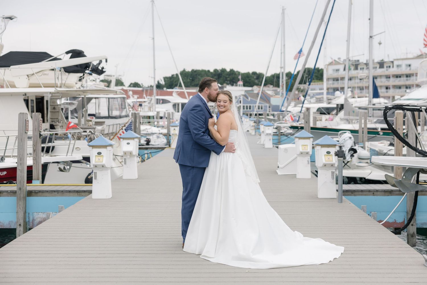 Bride and groom embrace on a marina dock surrounded by sailboats on their wedding day.