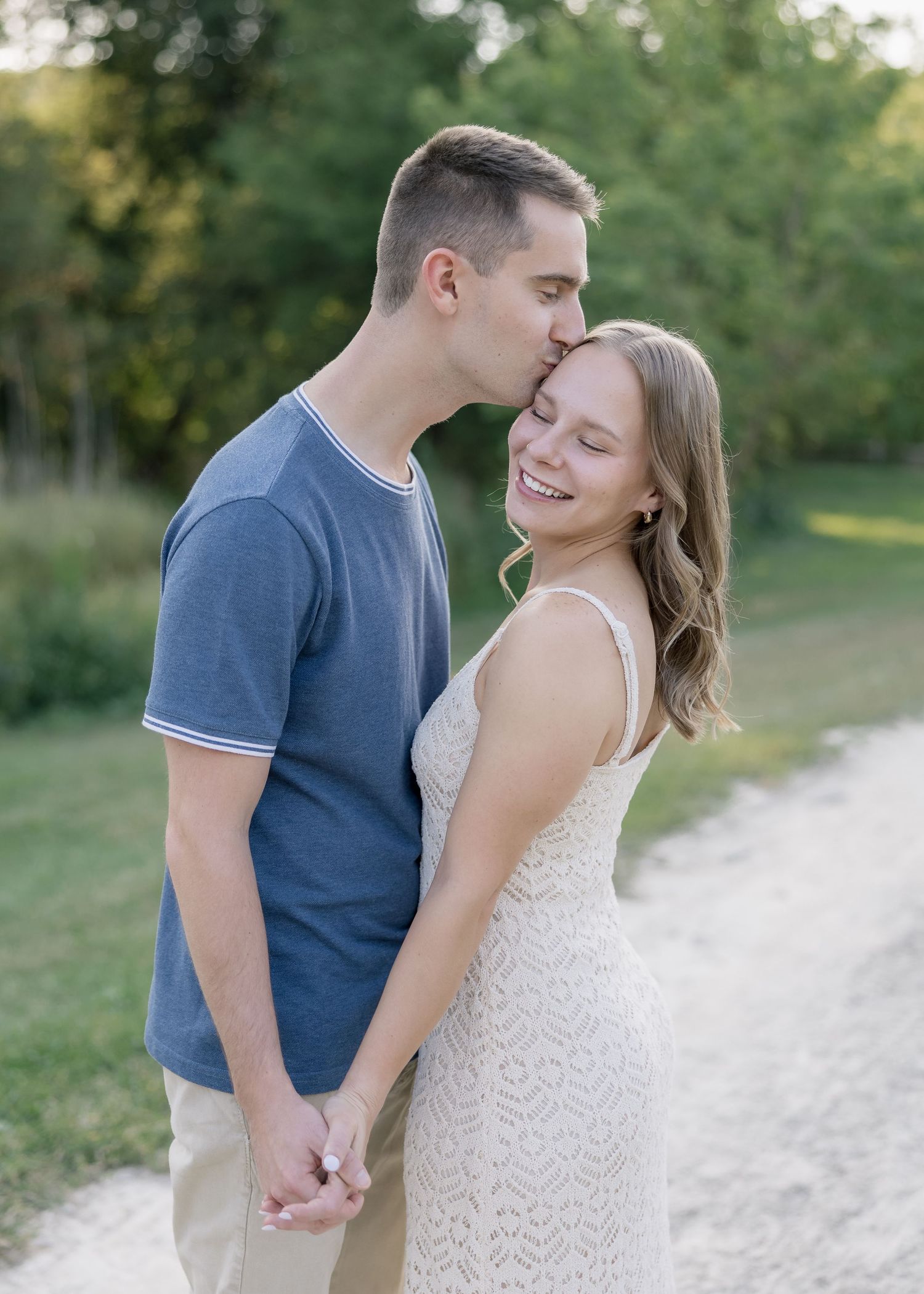 A romantic couple shares a tender moment on a dirt path surrounded by green trees during a summer photoshoot.