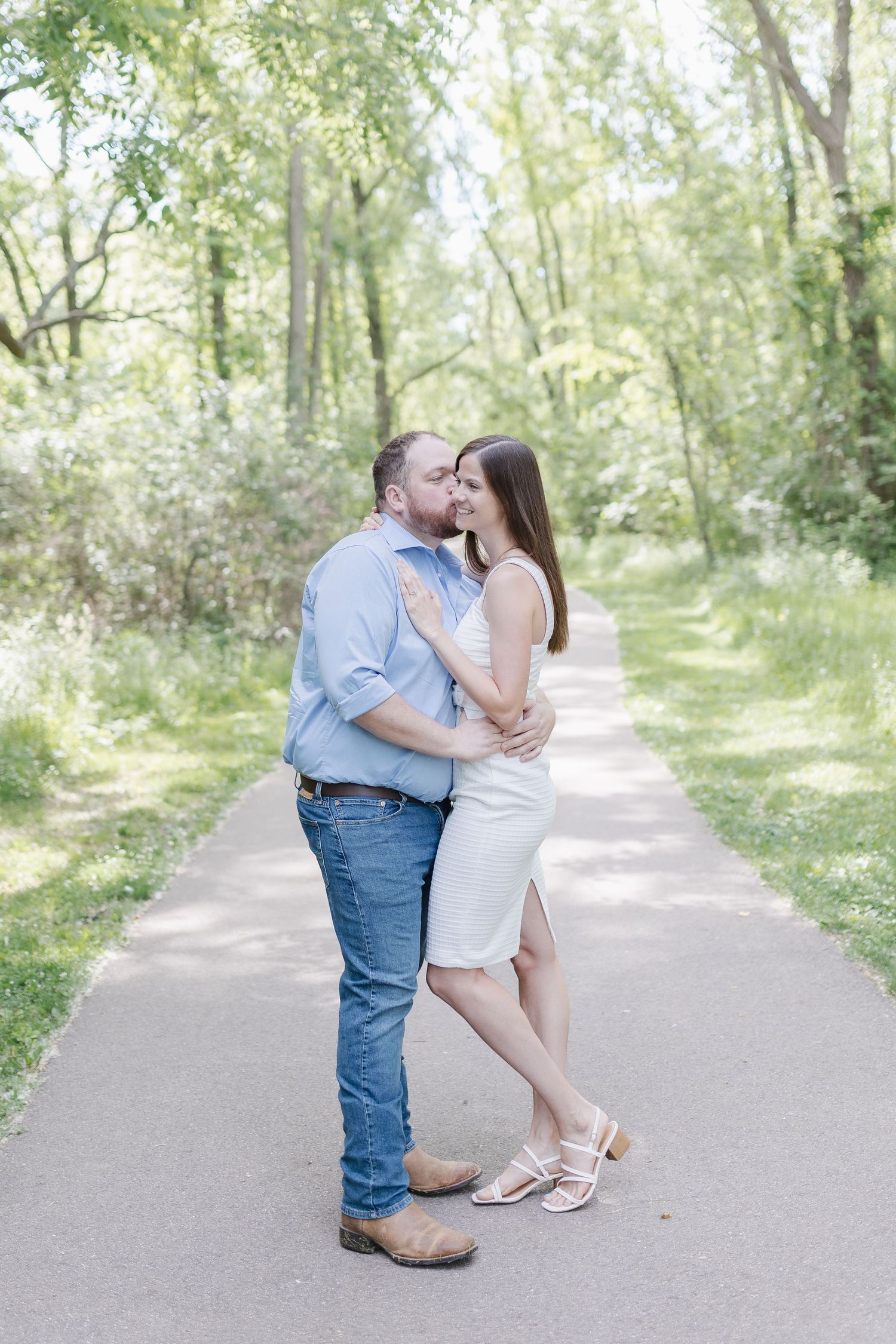 A romantic couple embraces on a sunlit nature path surrounded by lush green trees.