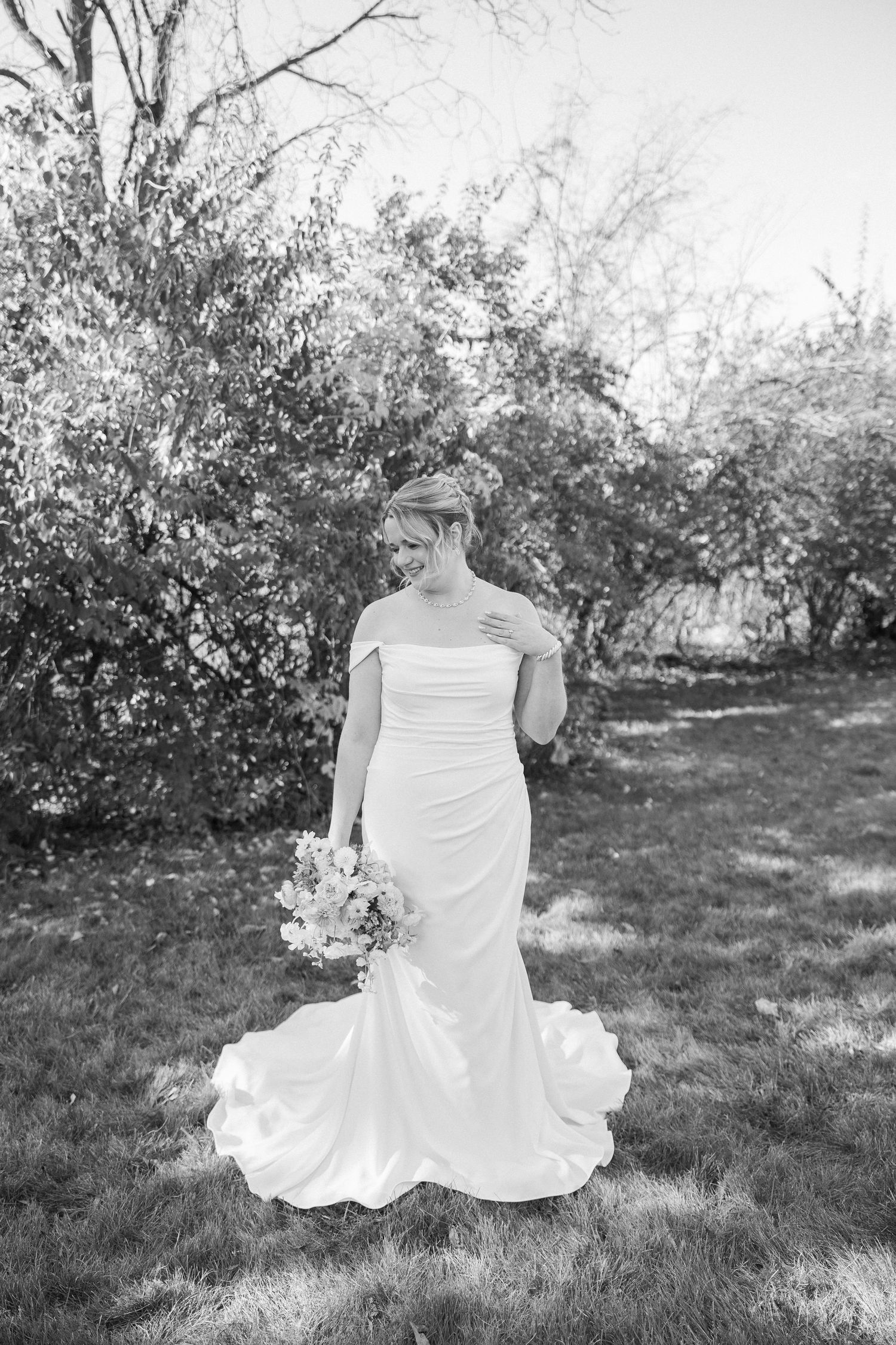 A bride in a fitted white wedding gown holds a bouquet while standing in a garden setting, captured in black and white.