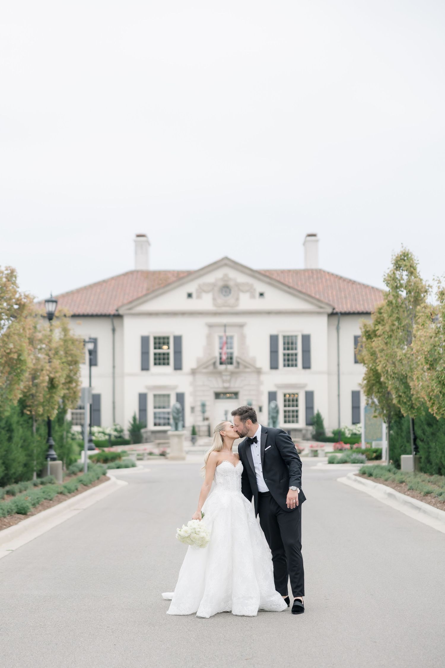 Bride and groom share a romantic kiss on a grand estate driveway, bride in strapless gown holding white bouquet.