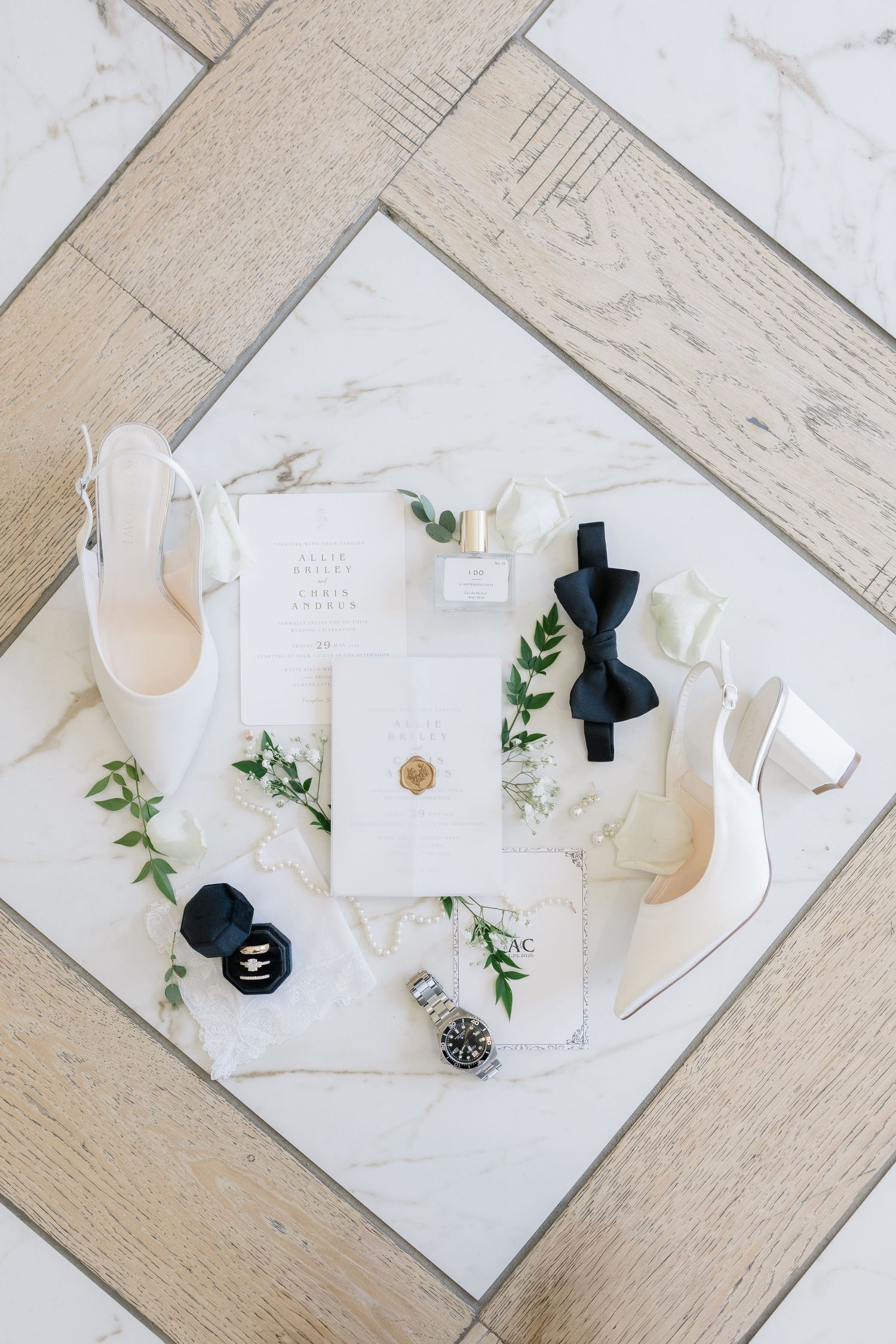 Elegant wedding flat lay with white heels, black bow tie, invitation suite, perfume, rings, and greenery on marble.