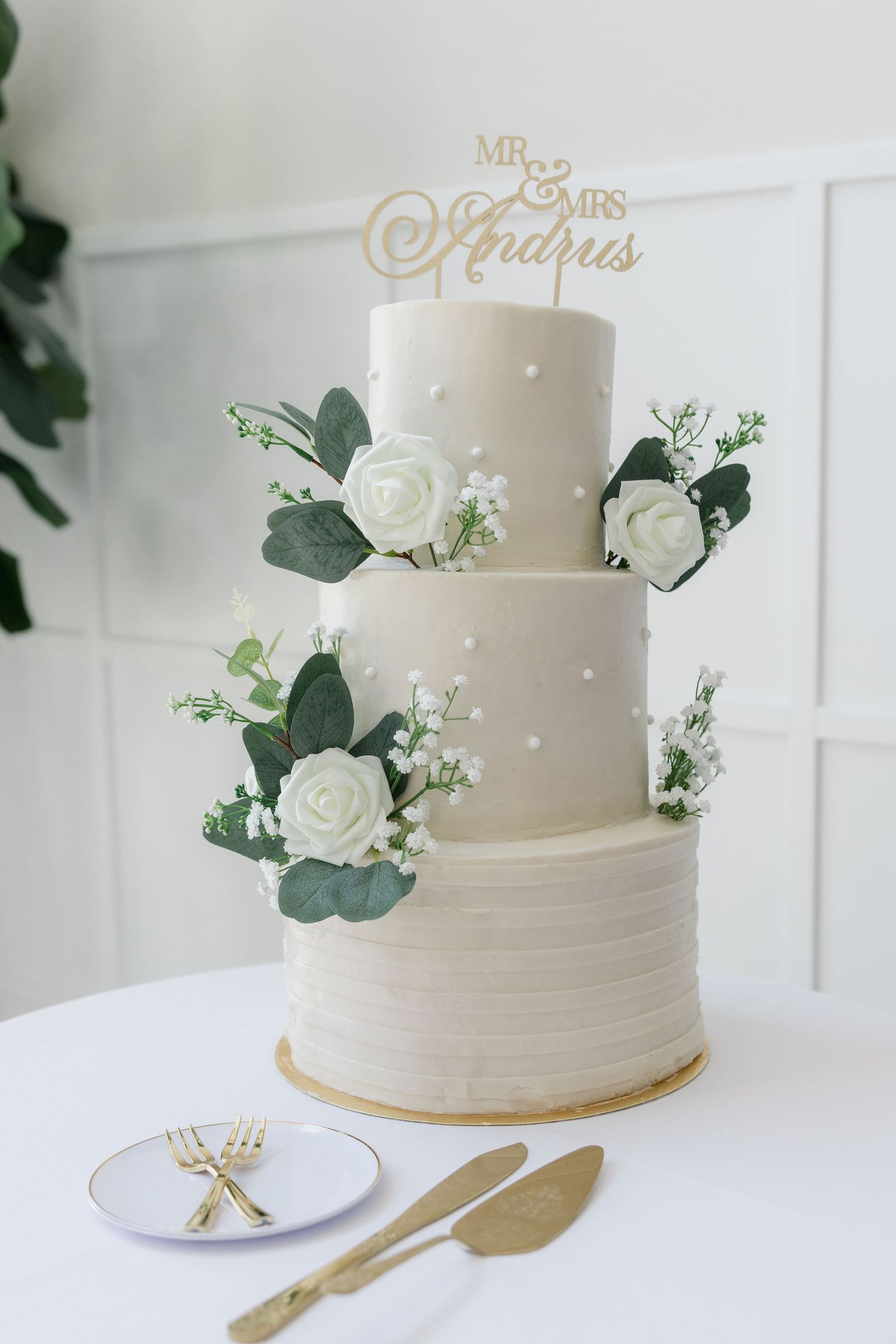 Elegant three-tier white wedding cake with green eucalyptus, white roses, and a gold Mr. & Mrs. Andrus cake topper.