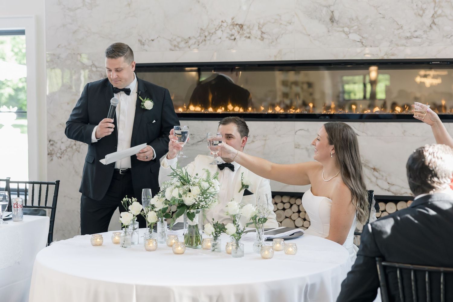 Groom giving a toast at wedding reception with bride raising champagne glass near floral centerpiece by elegant fireplace.
