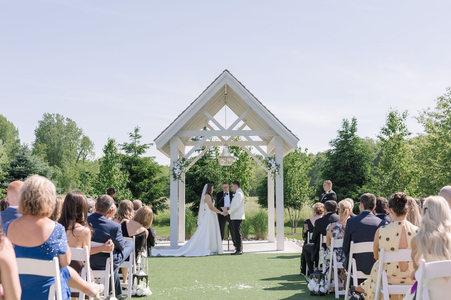 Outdoor wedding ceremony under white gazebo with guests seated on white chairs on a sunny day.