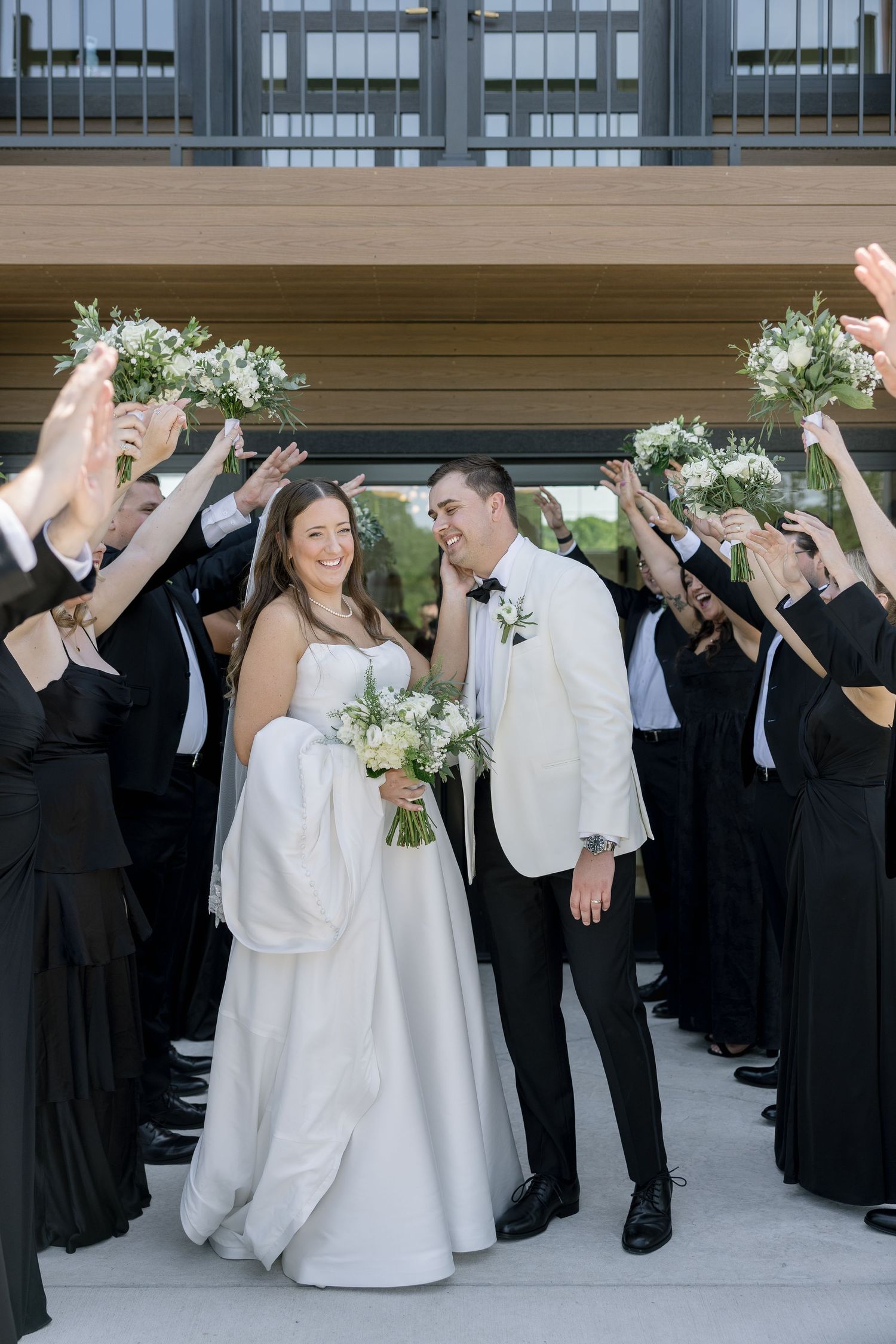 Bride and groom walk through arch of raised bouquets held by wedding party in black attire outdoors.