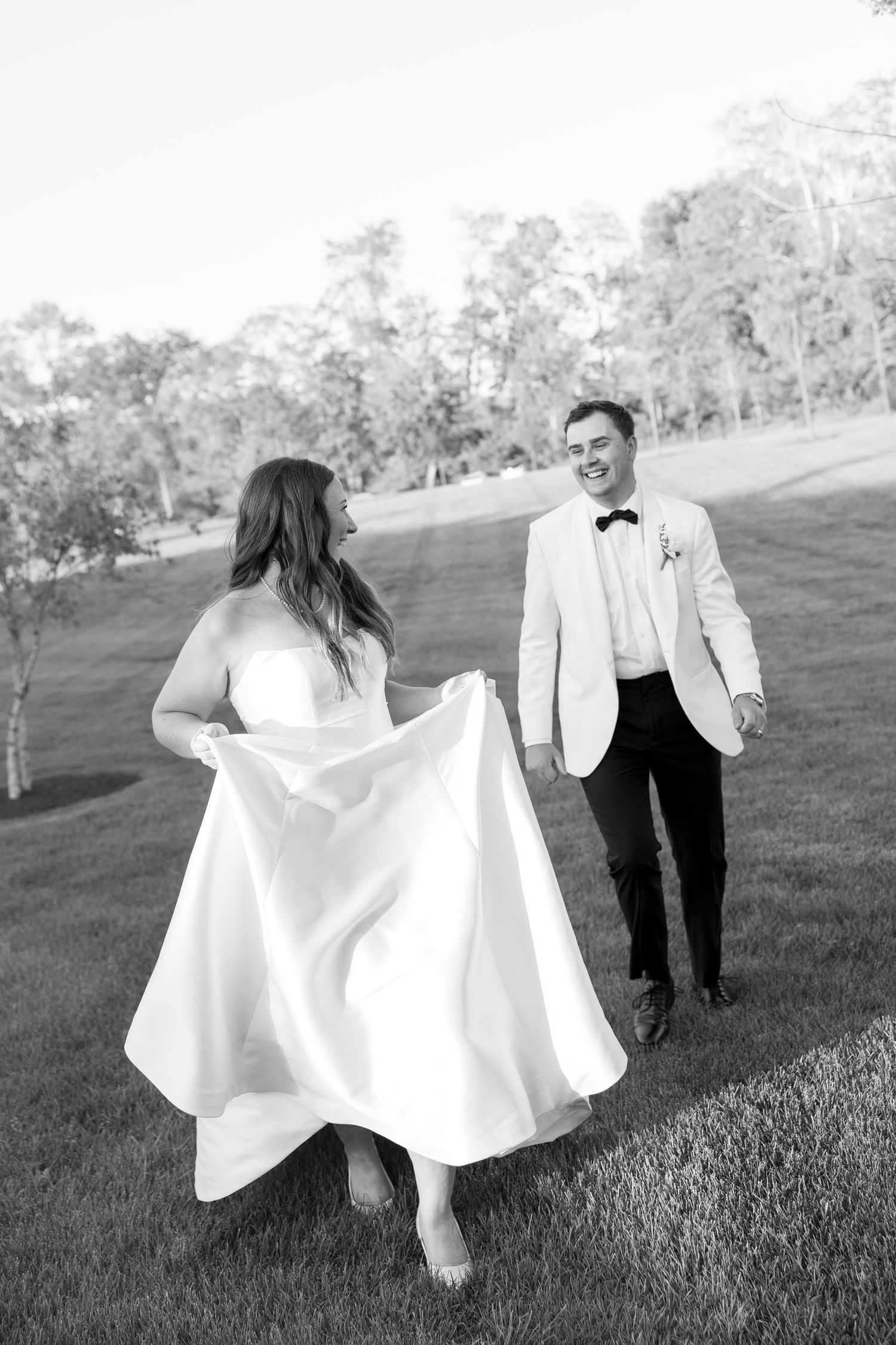 Bride and groom laugh while walking on green lawn, groom in white tuxedo jacket and black pants.