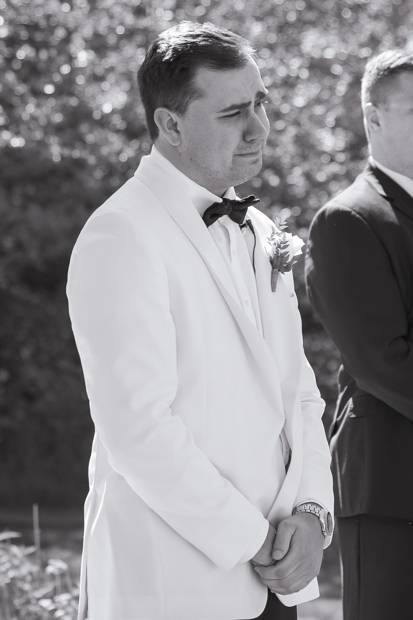 Black and white photo of a groom in white tuxedo and bow tie with boutonniere, emotional at outdoor wedding ceremony.