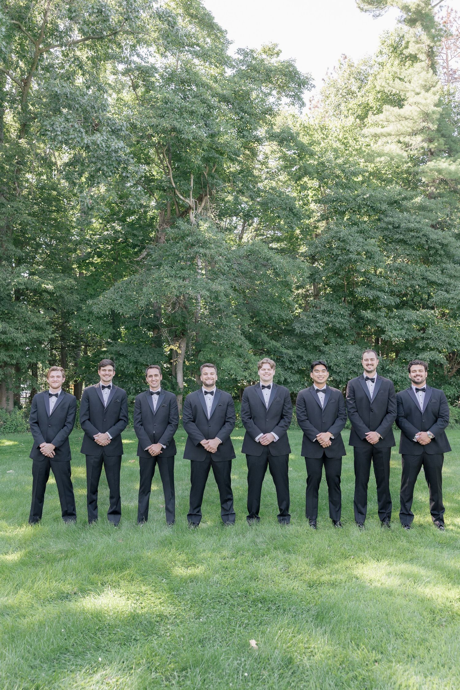 Groomsmen in black tuxedos and bow ties stand in a line formation on a grassy lawn surrounded by trees.