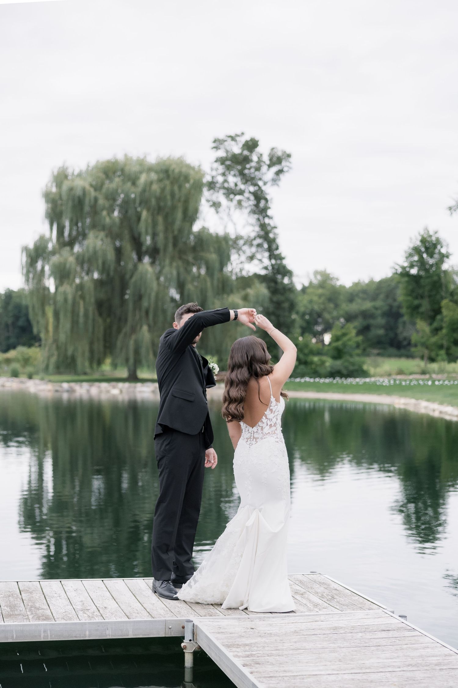 A couple shares a romantic dip pose on a wooden dock by a serene lake with willow trees in the background.