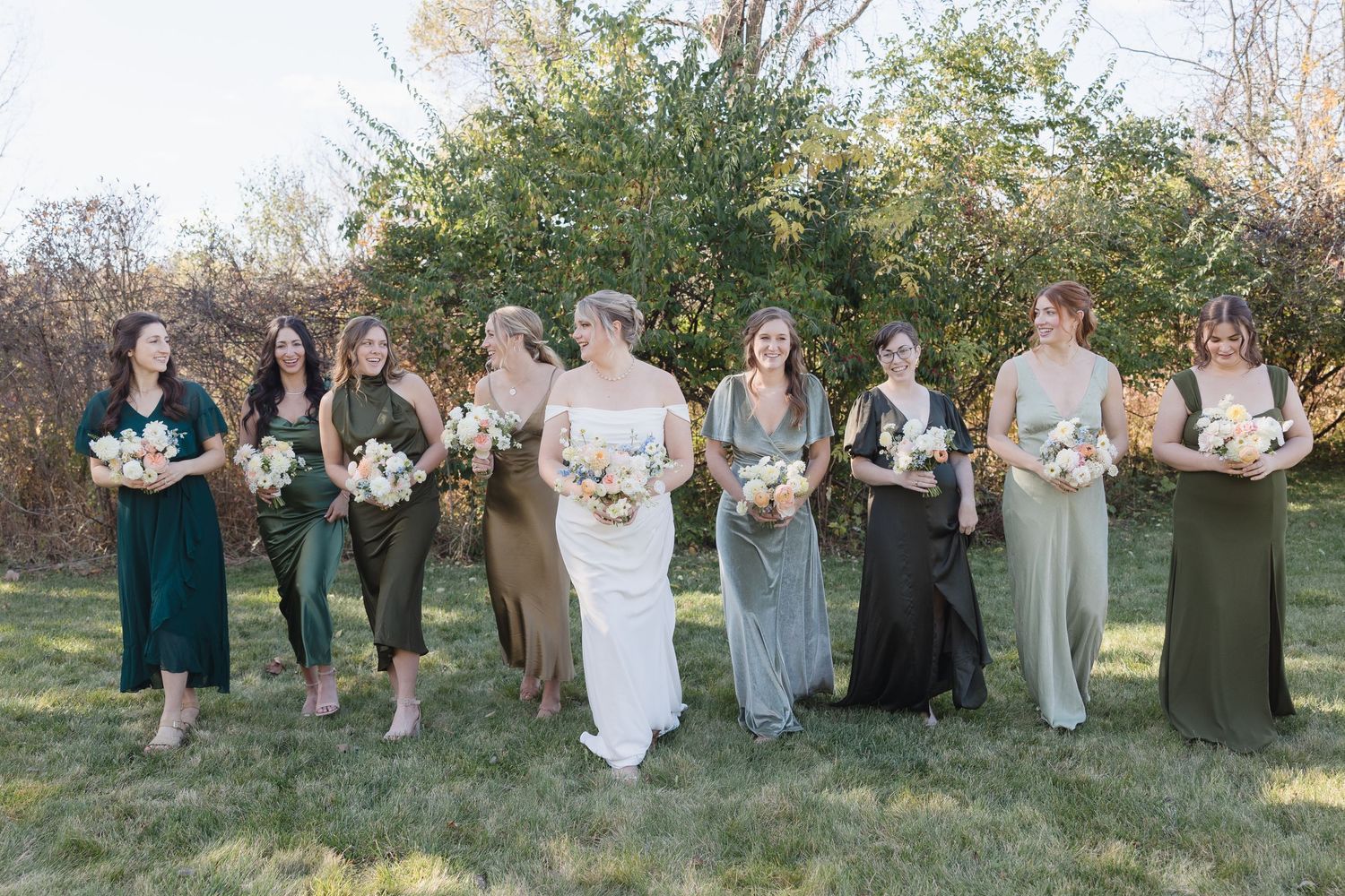Outdoor wedding party photo with bridesmaids in mixed green tones holding coordinated floral arrangements.