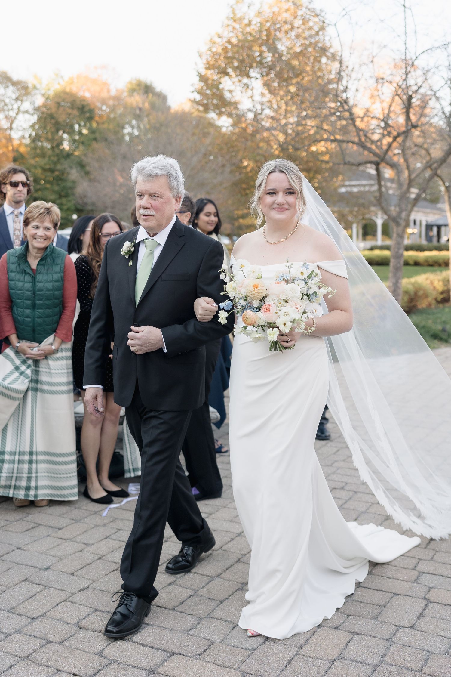 A wedding procession walks down a brick pathway surrounded by autumn scenery.