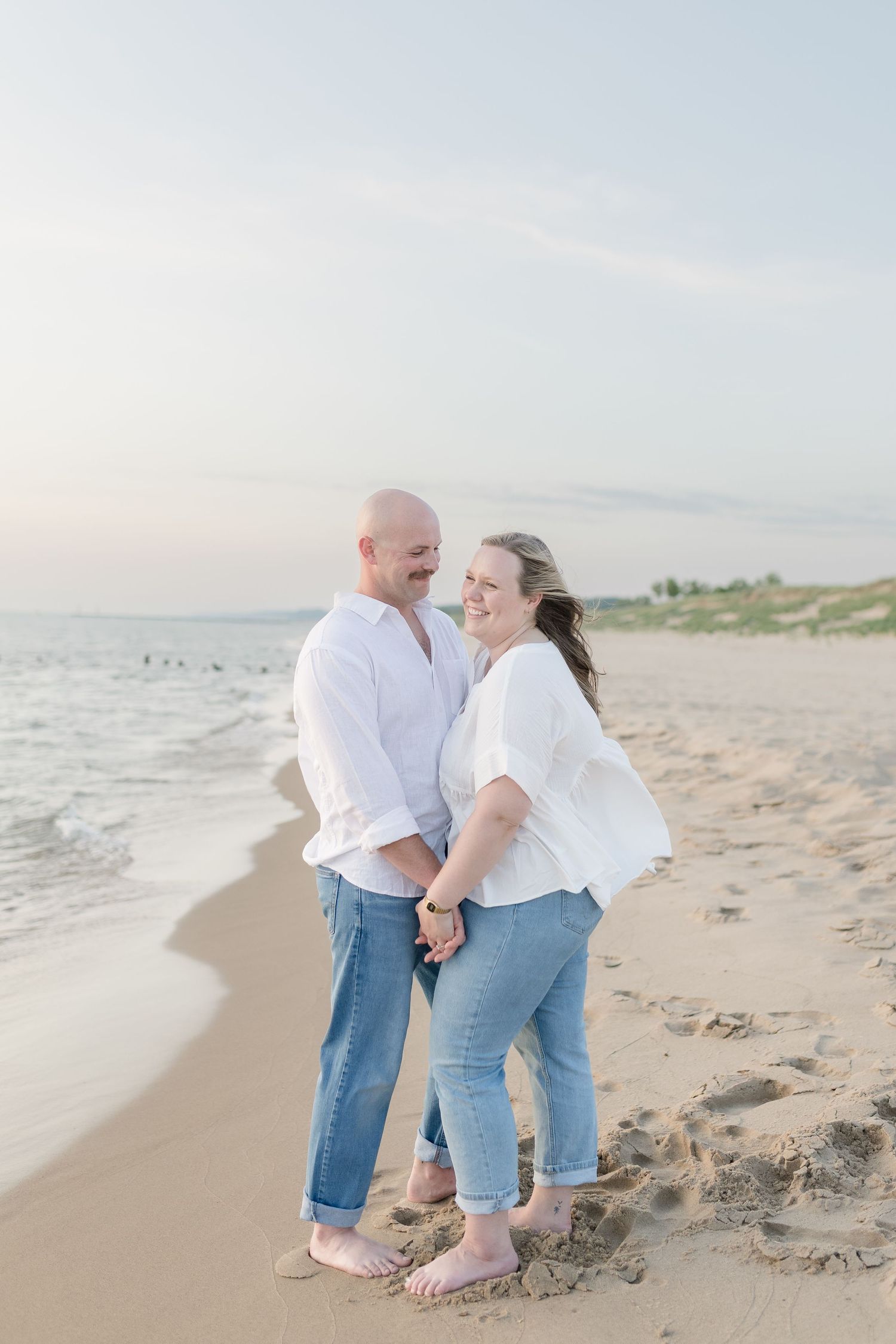 A couple in matching white shirts and jeans walks barefoot along a serene beach at sunset.