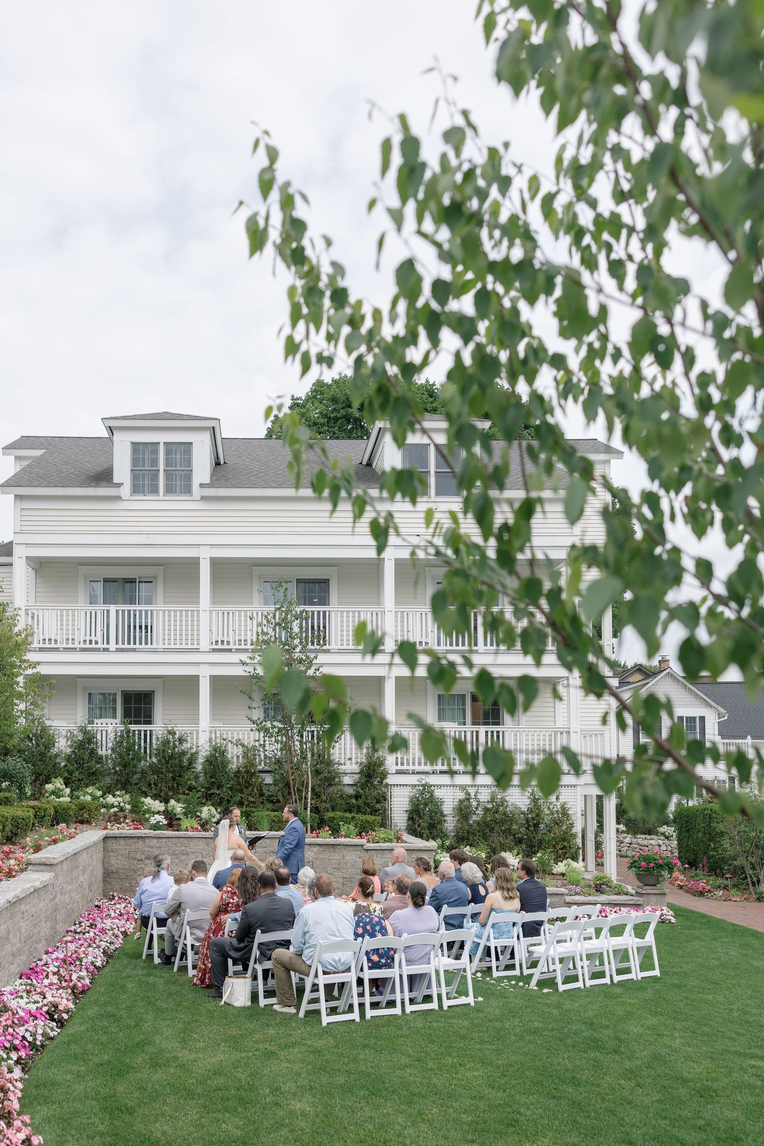 Outdoor wedding ceremony on a green lawn with white chairs in front of a large white colonial-style venue building.