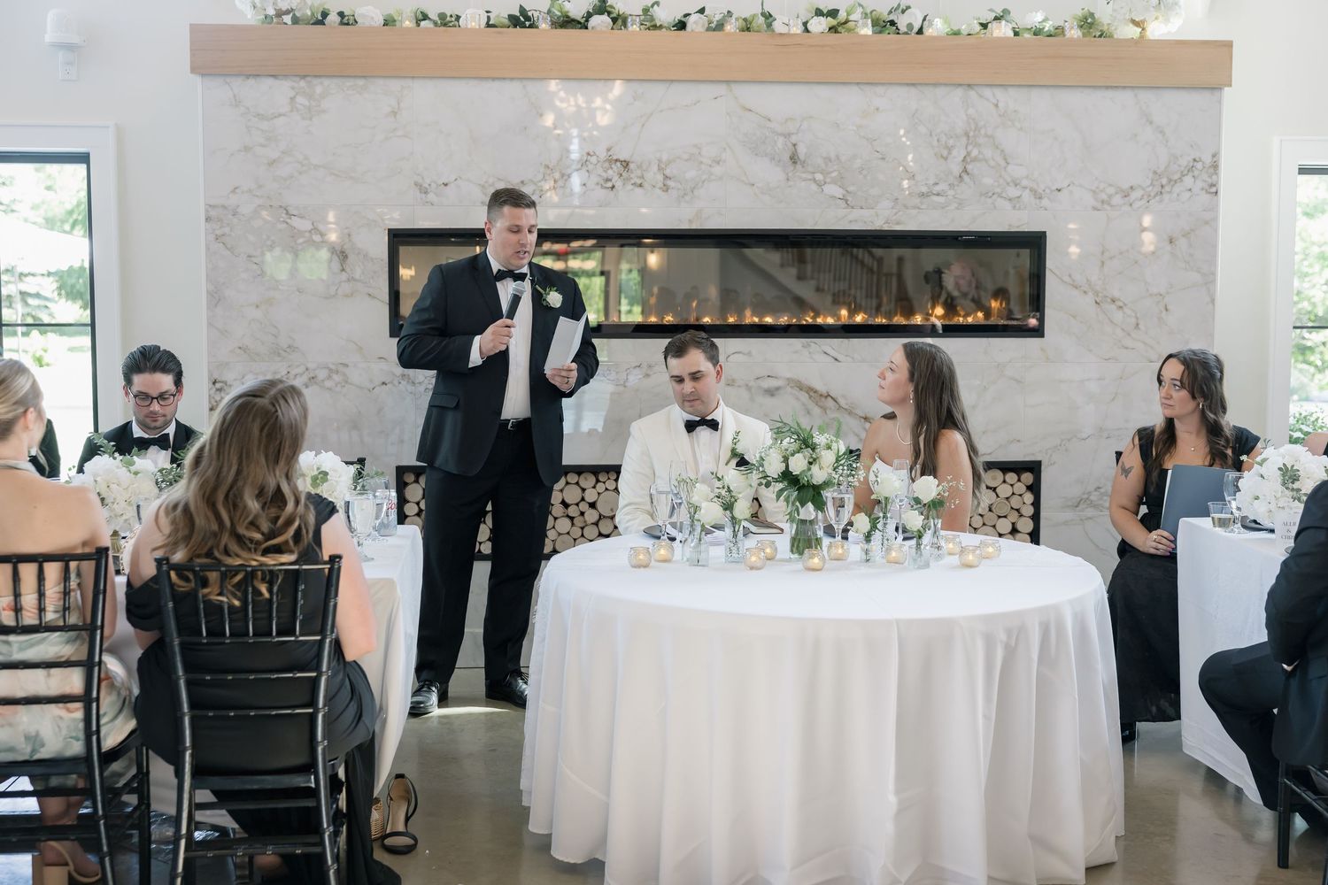 Man in tuxedo gives wedding toast at elegant reception with fireplace backdrop while bride and groom sit at sweetheart table.