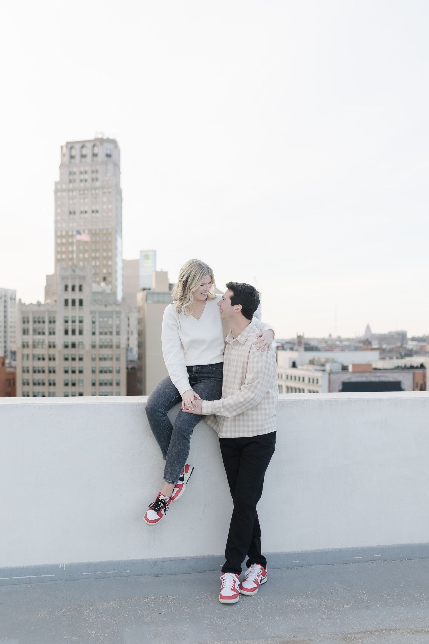 Couple sitting on rooftop ledge with urban skyline in background wearing casual outfits and matching sneakers.