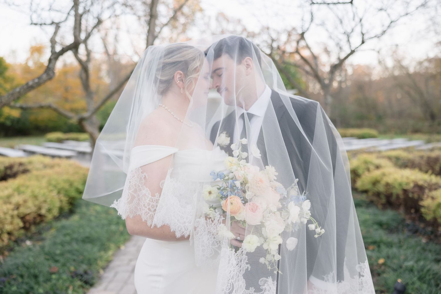 A couple shares an intimate moment under a flowing veil while holding a pastel floral bouquet in an autumn garden.