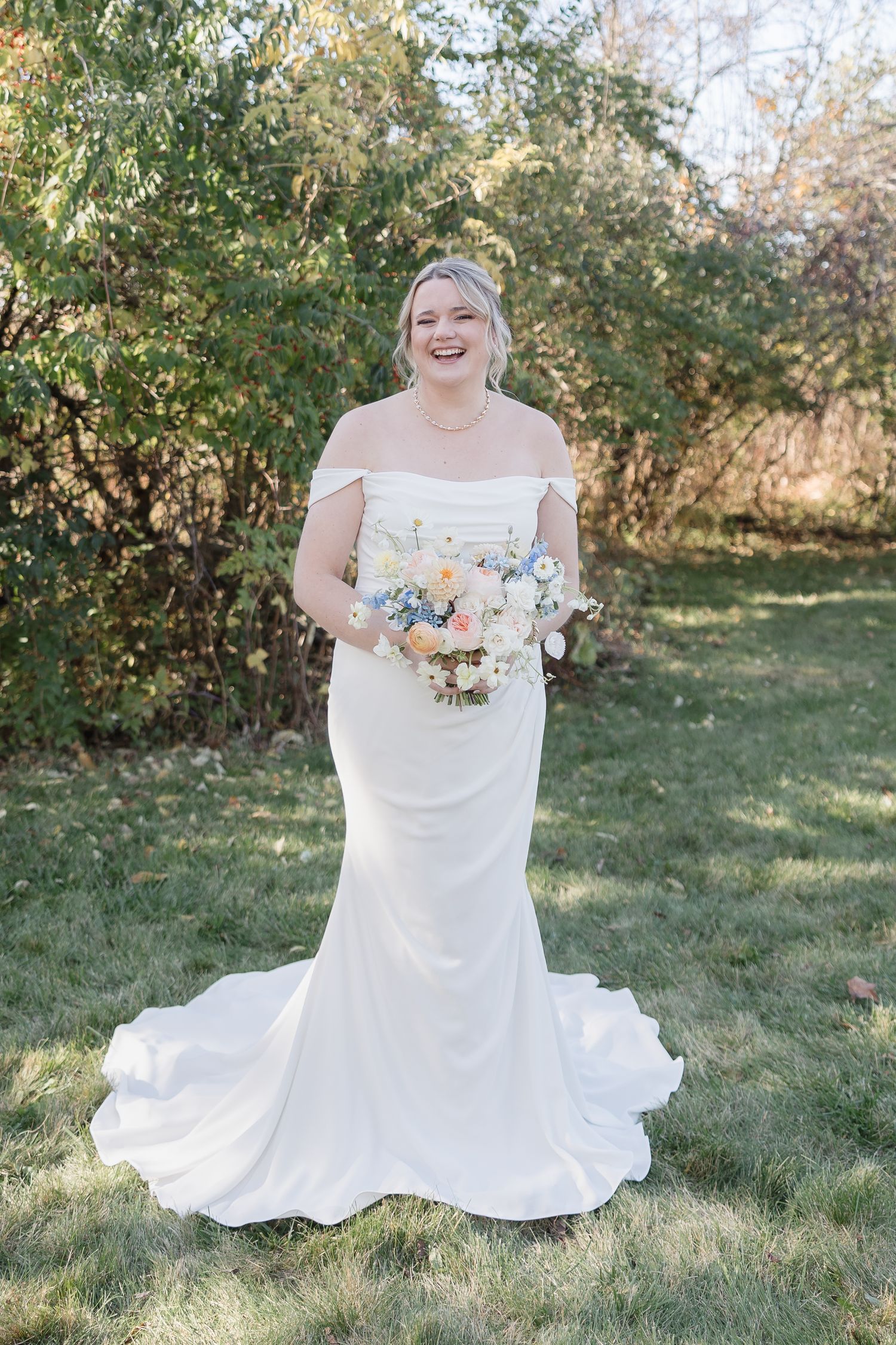 A bride in an elegant off-shoulder white mermaid wedding gown holds a pastel bouquet while standing in a garden setting.