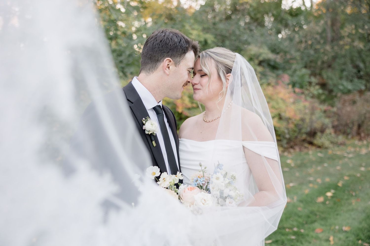 Romantic wedding couple shares an intimate moment outdoors with flowing veil and autumn foliage in background.