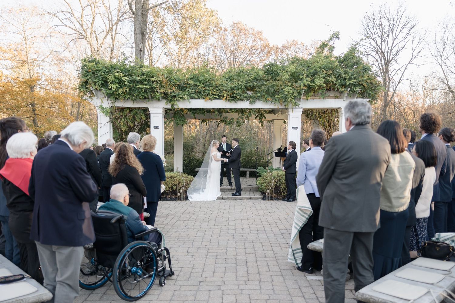 Wedding ceremony setup with white pergola and guests seated on paved courtyard.