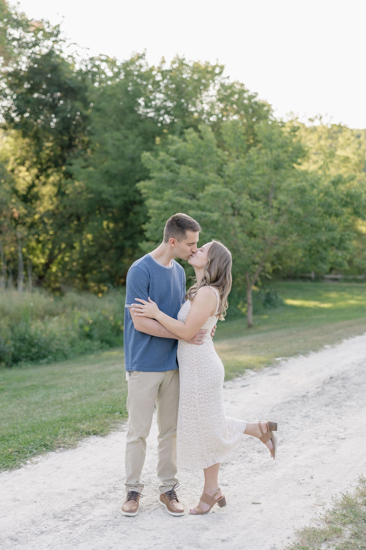 A couple shares a romantic kiss on a sunlit dirt path surrounded by lush green trees and foliage.