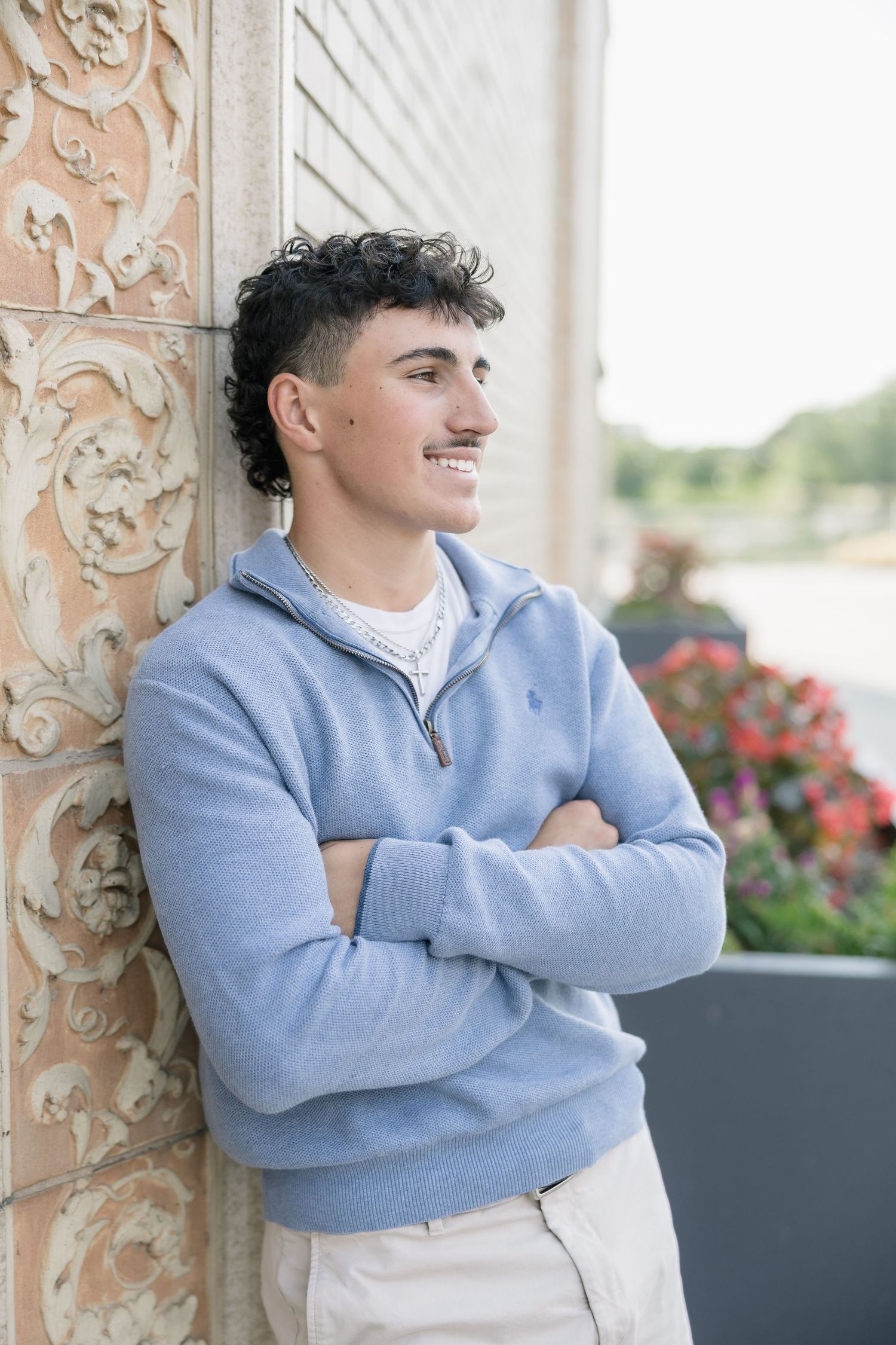 A person wearing a light blue sweater leans against an ornate column with decorative carvings and flowers in the background.