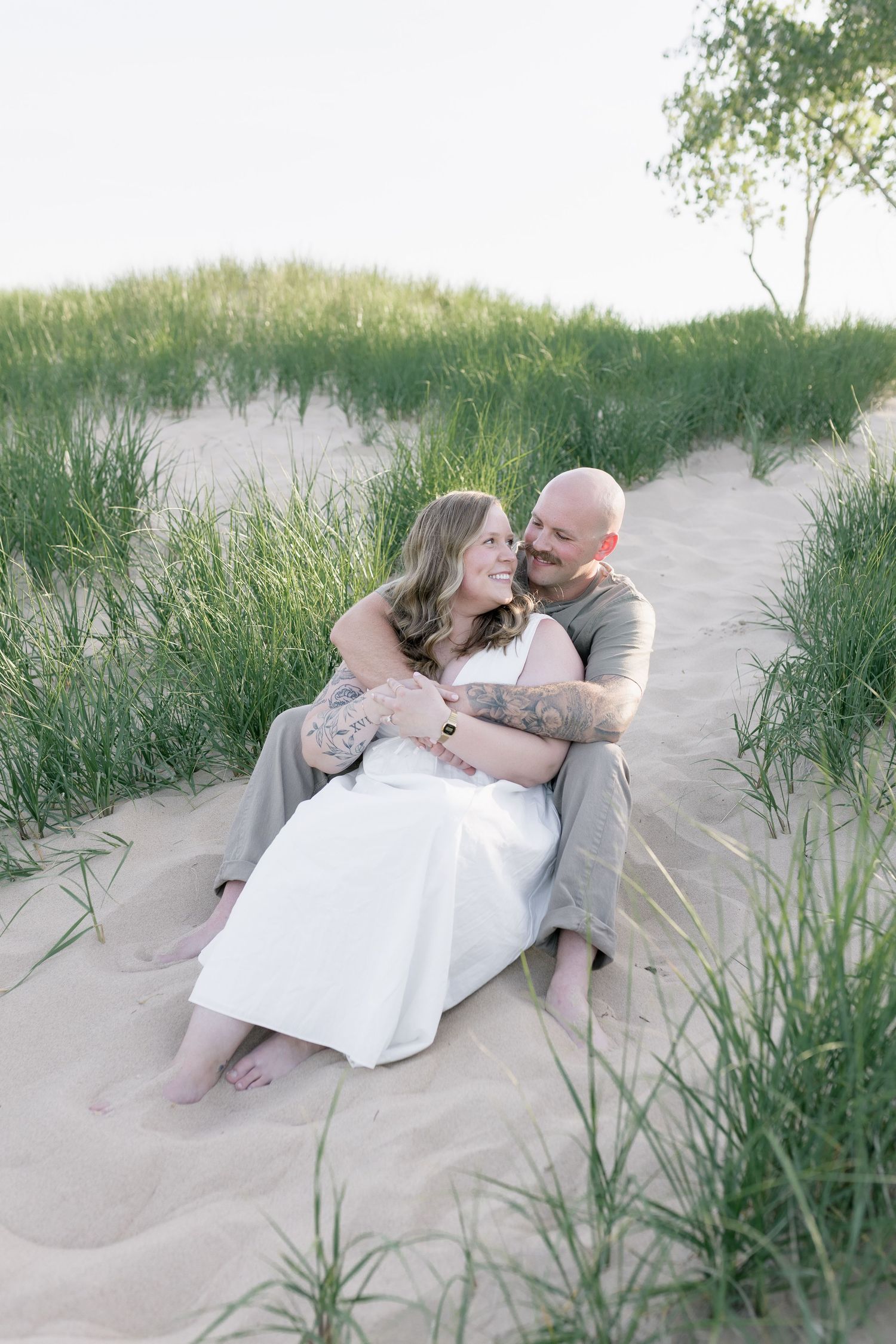 A couple shares an intimate moment while lying on sandy beach dunes surrounded by tall grass at sunset.