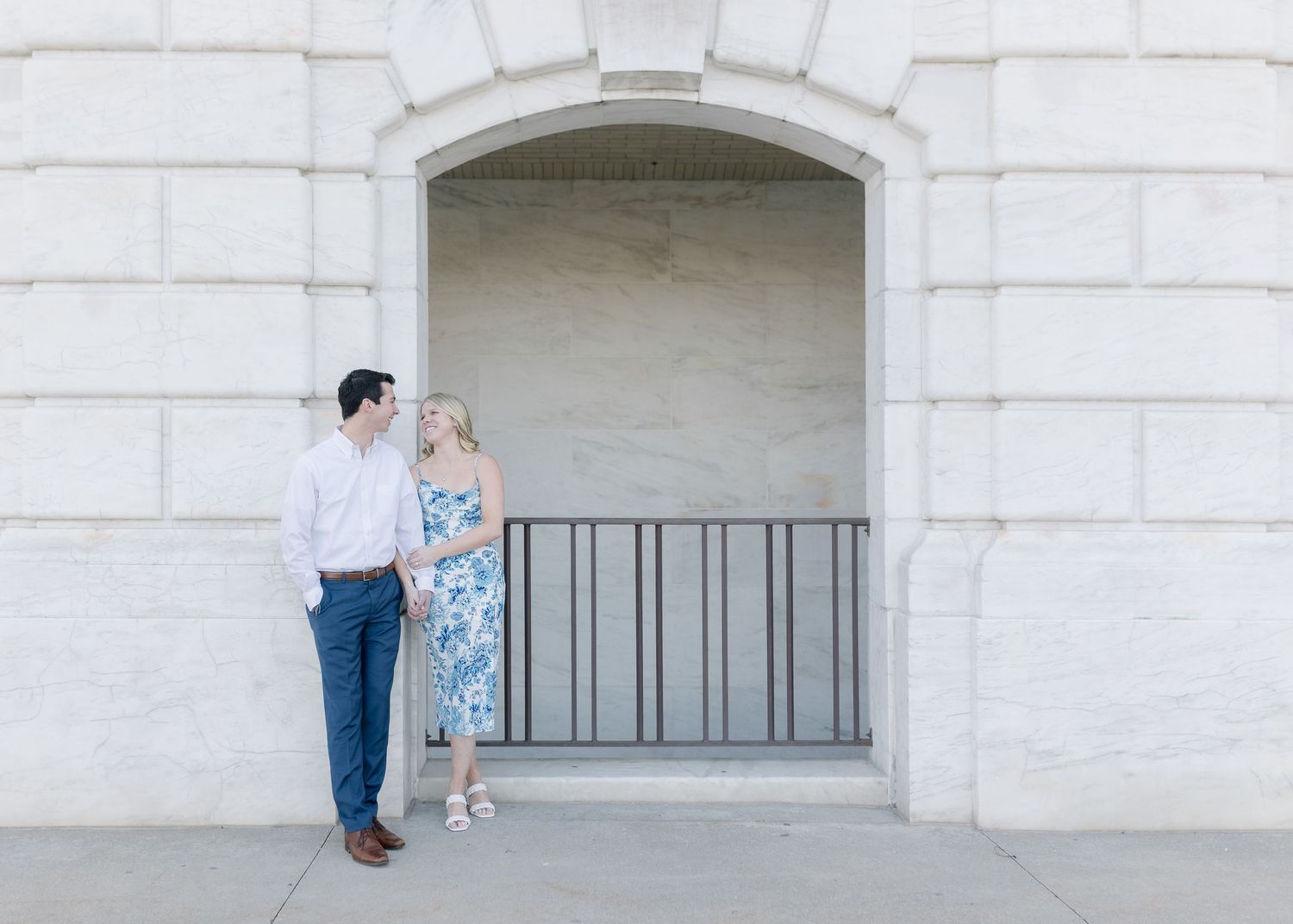 A couple stands together in an elegant stone archway with white walls and black iron railing on a bright sunny day.