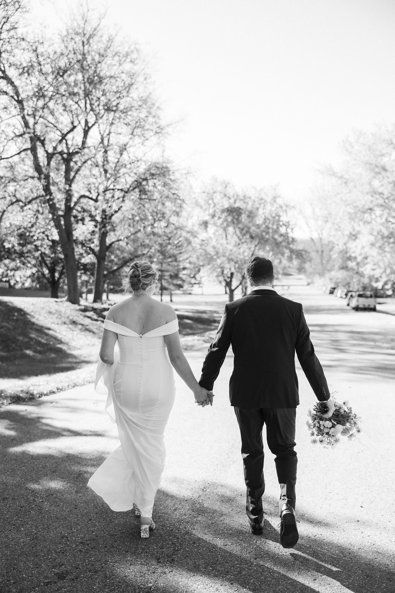 Black and white shot of couple walking hand in hand down tree-lined path after ceremony.