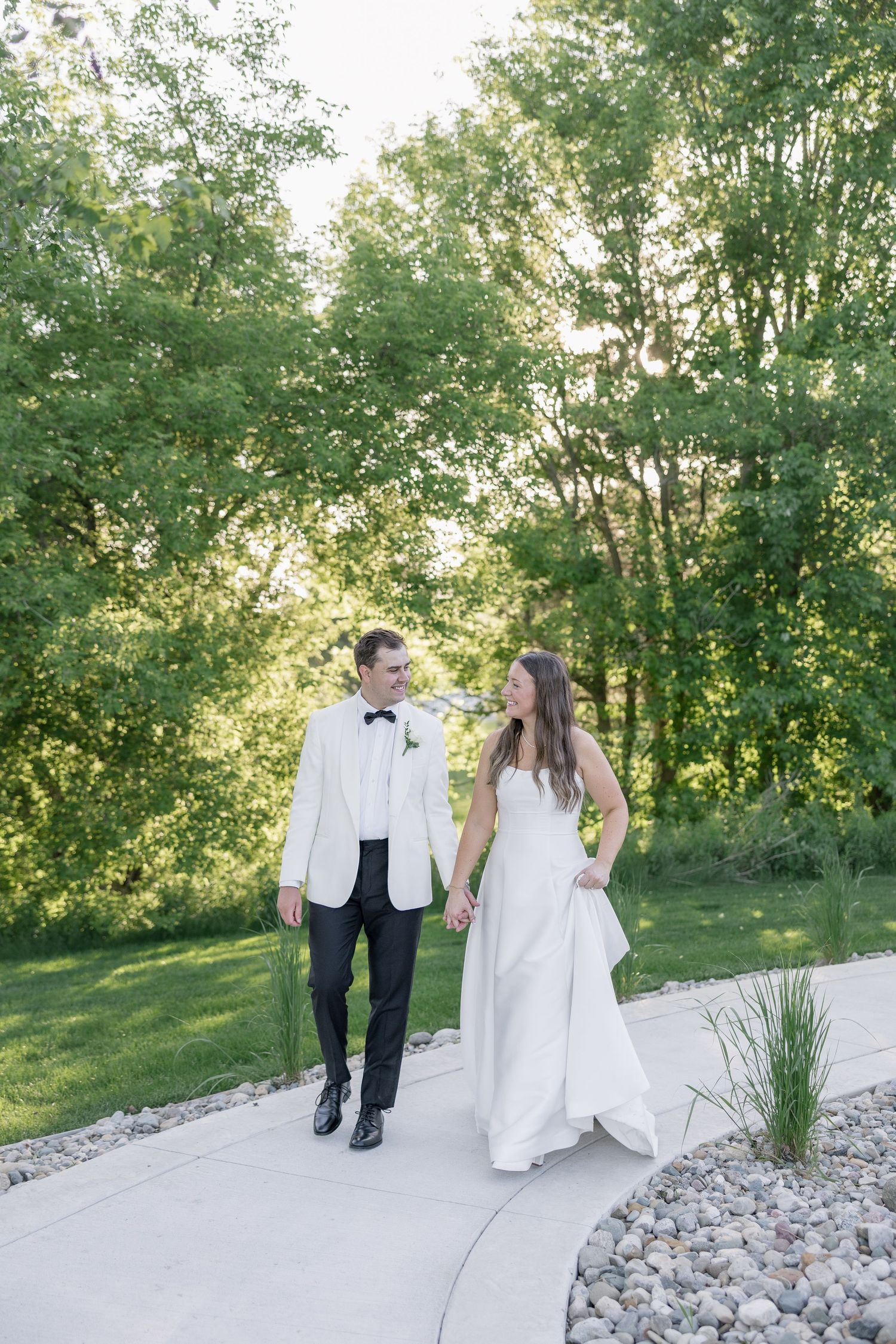 Bride and groom walk hand-in-hand on a garden path, smiling at each other in wedding attire surrounded by lush green trees.