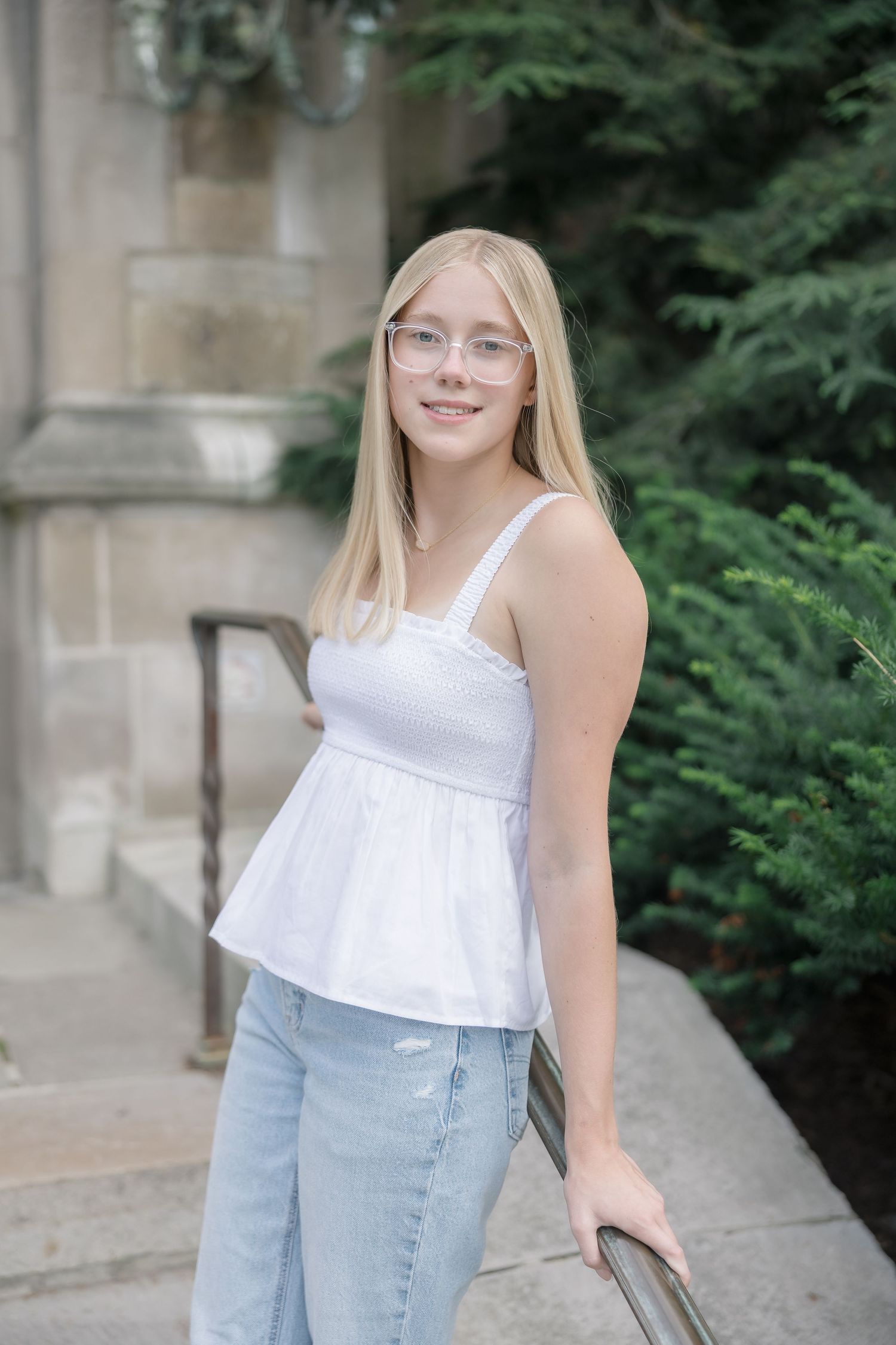 Young blonde woman in white top and jeans smiling outdoors near stone building and evergreen trees.