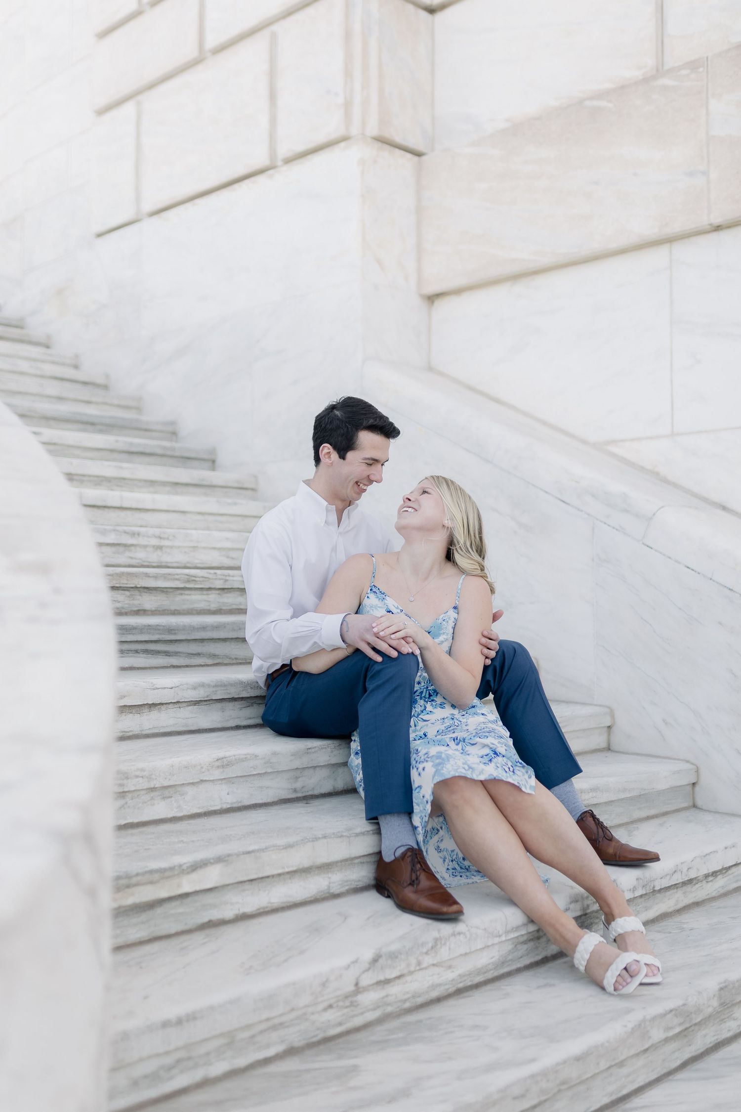 A couple sits intimately on white marble steps wearing casual summer attire and sharing a romantic moment.