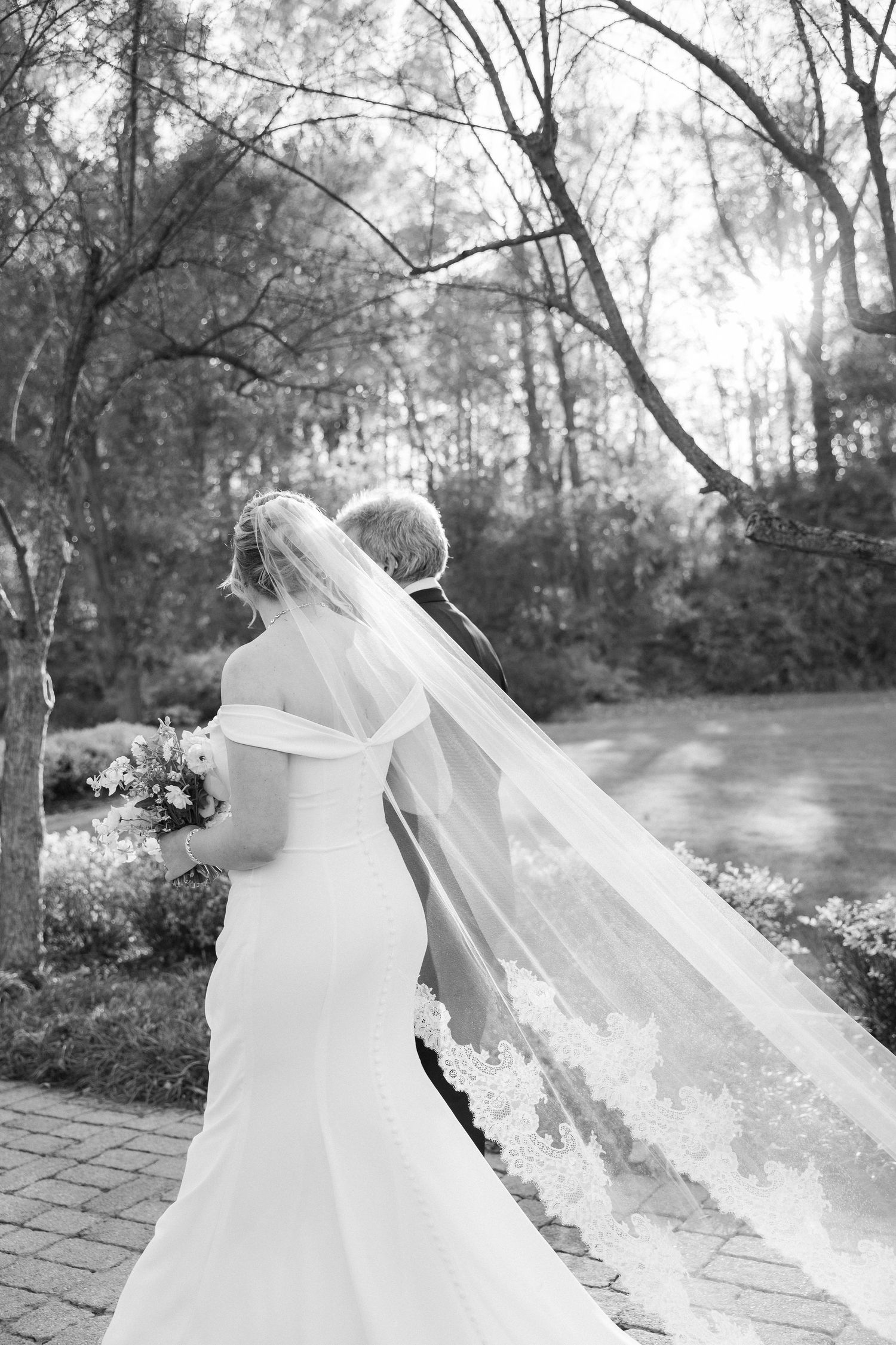 Bride in flowing white dress with long veil caught in breeze on stone pathway.
