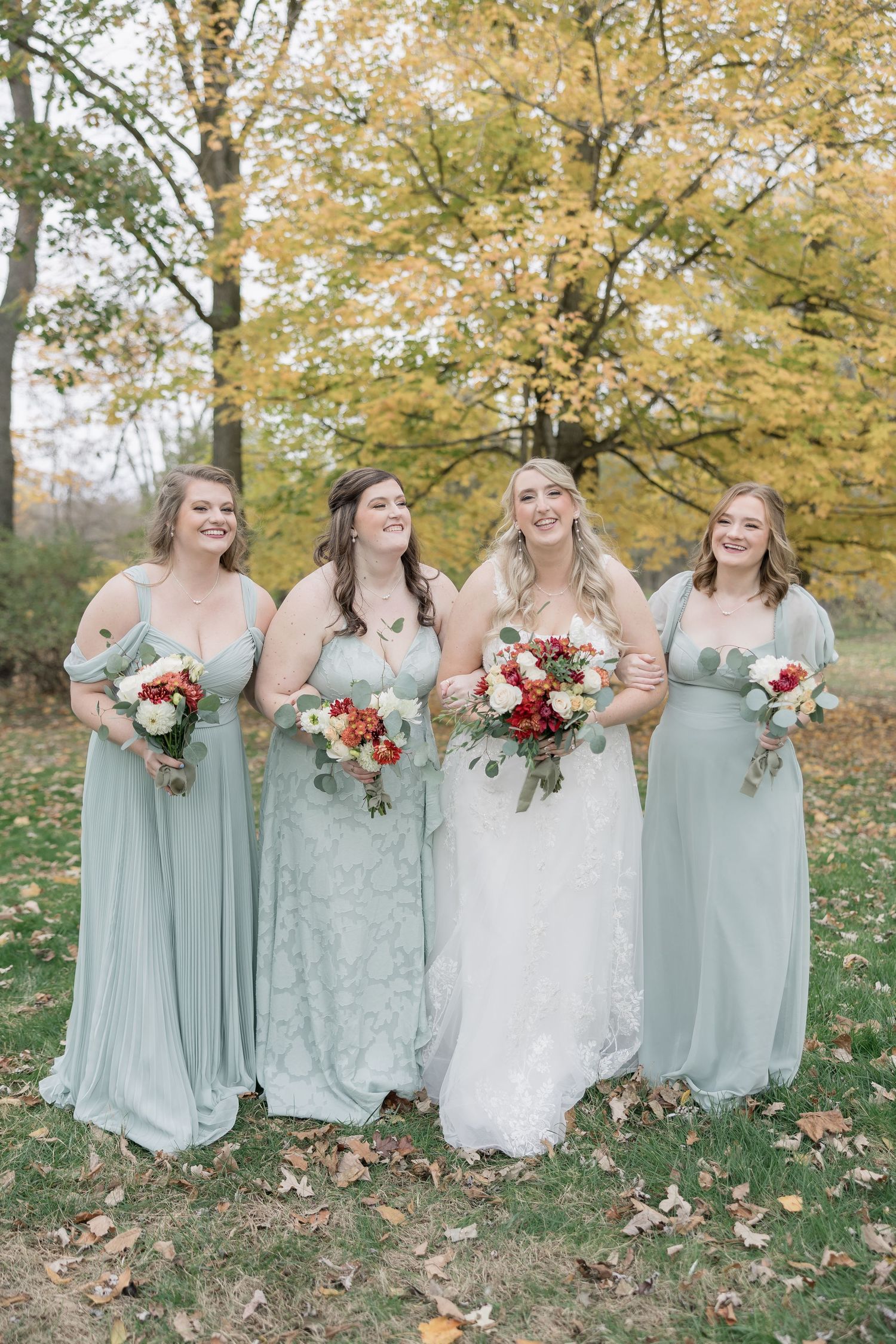 Bridal party in sage green dresses poses together on autumn day with fall foliage and bouquets.