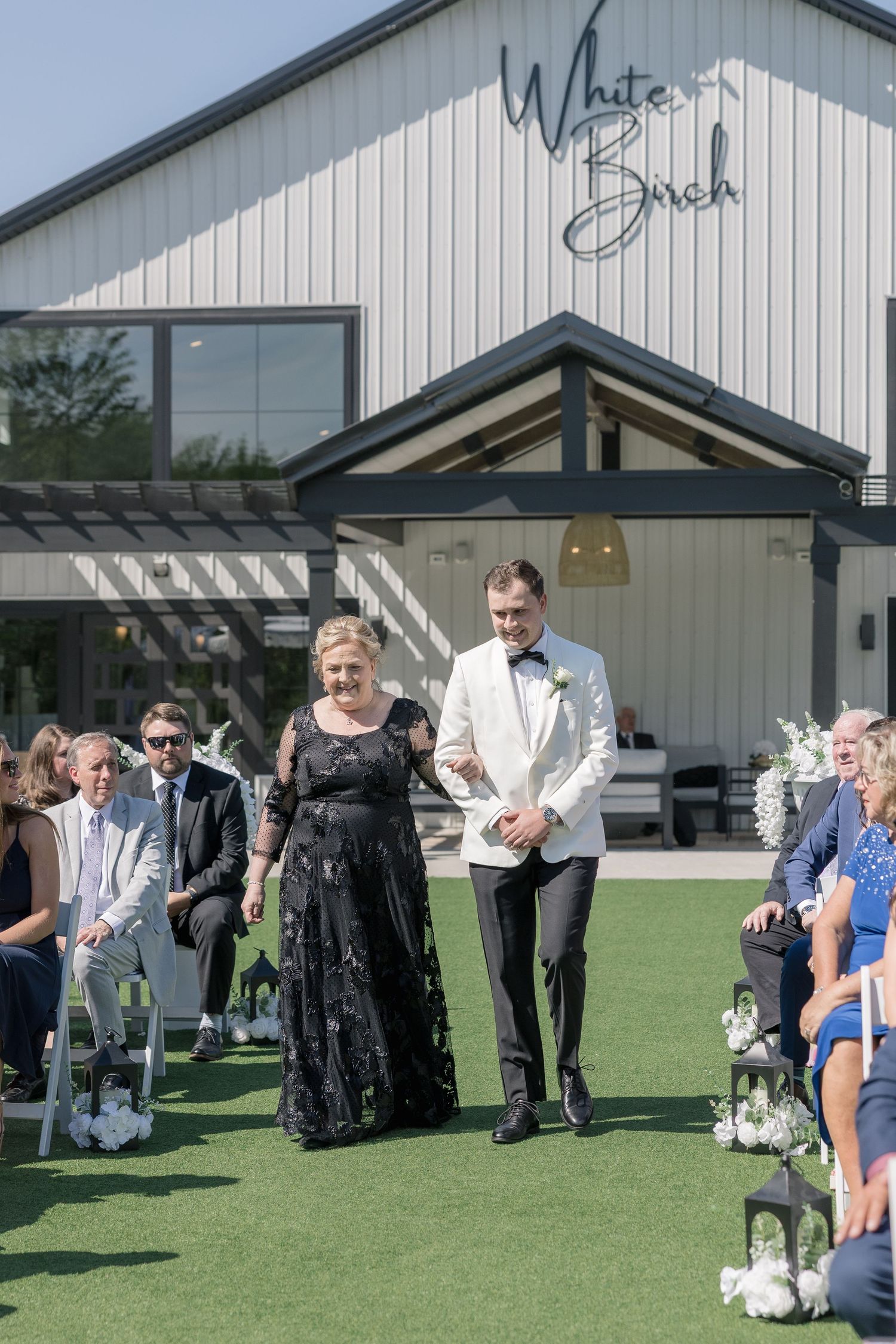 Groom in white tuxedo walks down aisle with woman at White Birch wedding venue outdoor ceremony.
