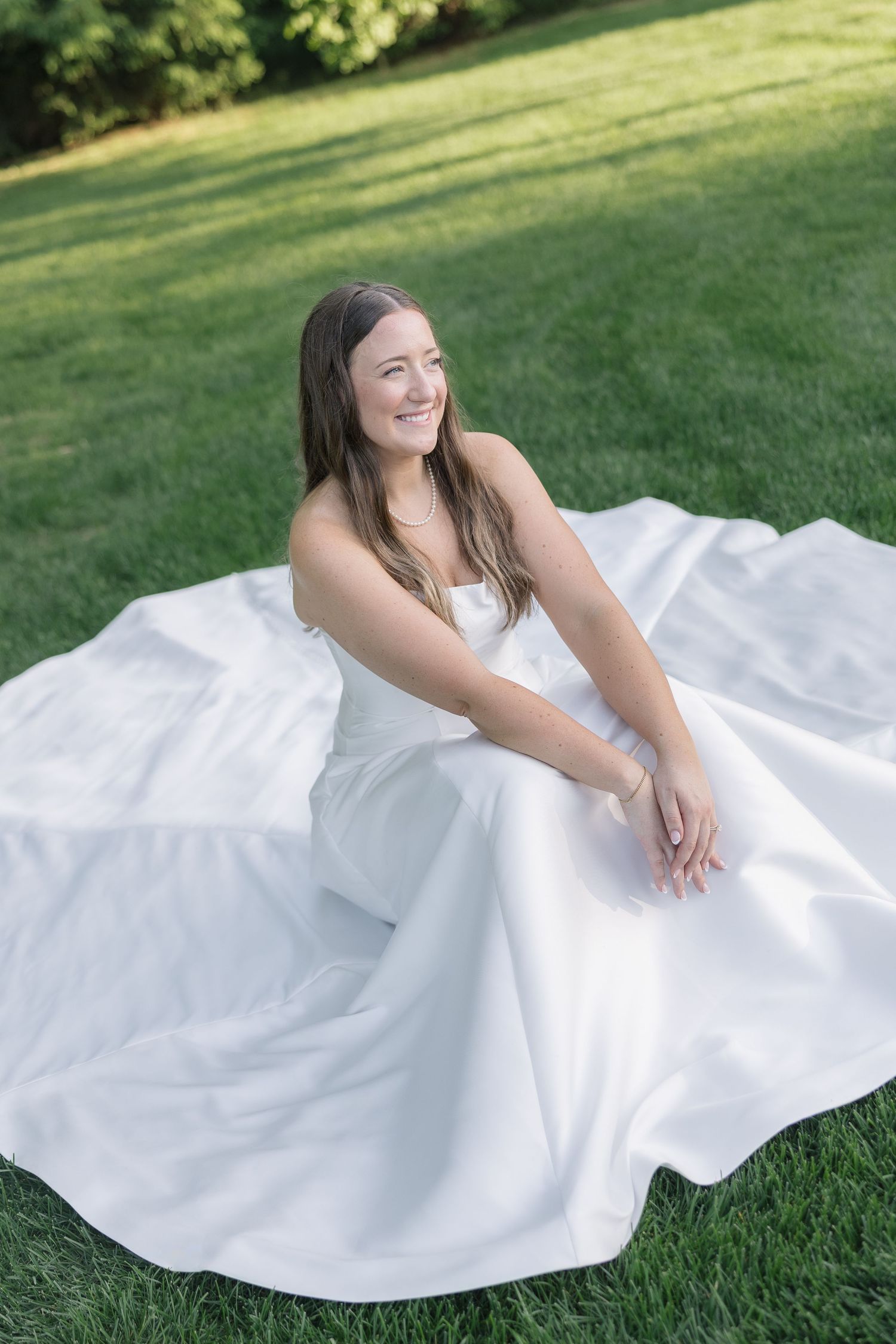 Smiling bride in elegant white wedding gown sitting on lush green grass, dress fanned out around her.