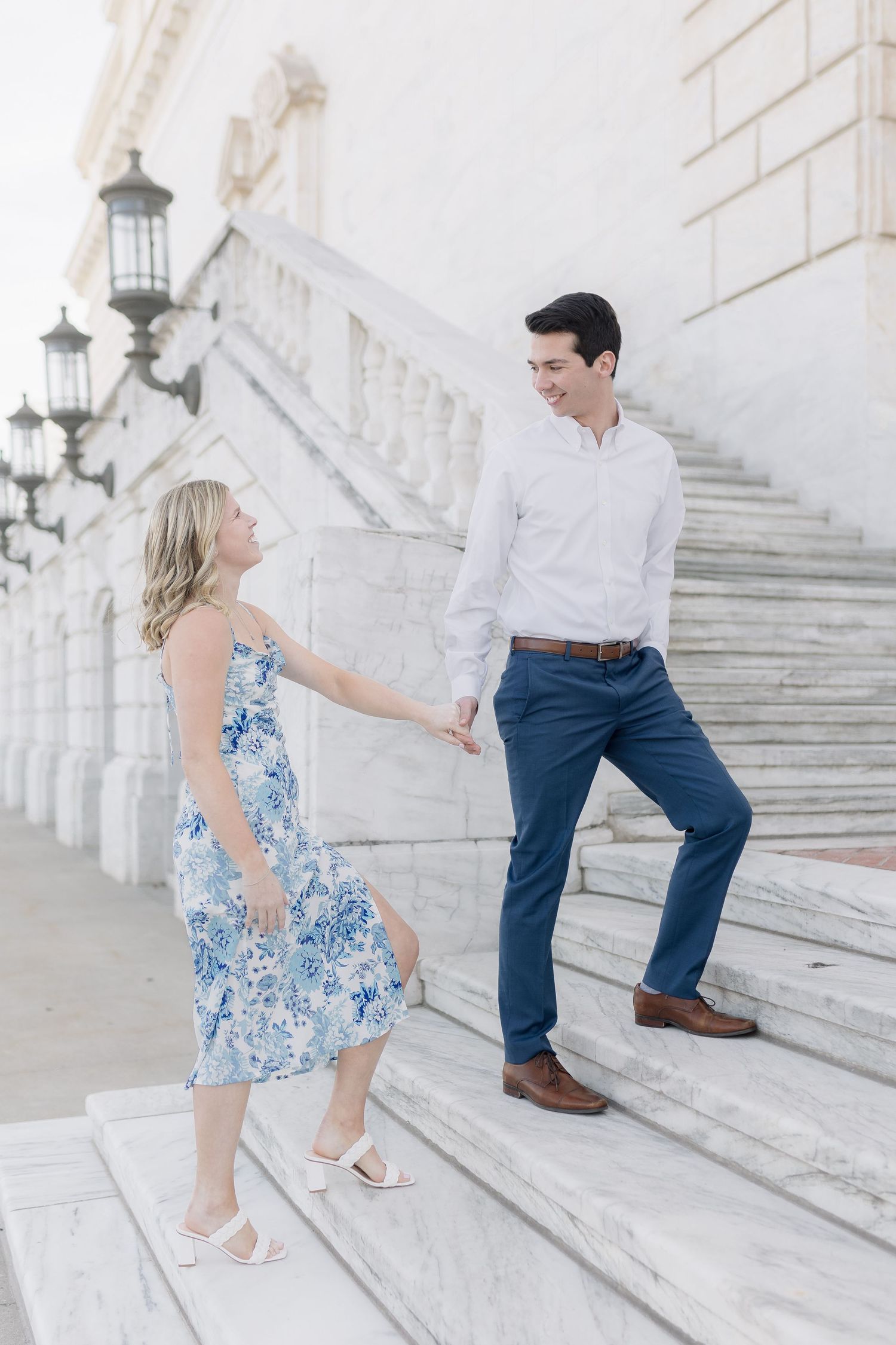 A couple on white marble steps, with her in a blue floral dress and him in a white shirt and jeans.